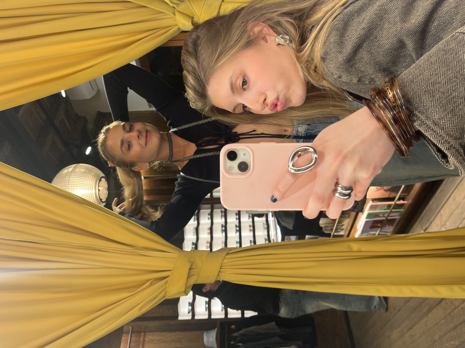 A blonde woman takes a mirror selfie with her pink phone. She is posing with puckered lips. Her outfit includes a gray blazer, jeans, and layered bracelets and rings. Her reflection is visible in the mirror, showing a second woman with a ponytail in a black top and choker necklace. The mirror is framed by thick yellow curtains. A lamp and clothing racks are visible in the background.