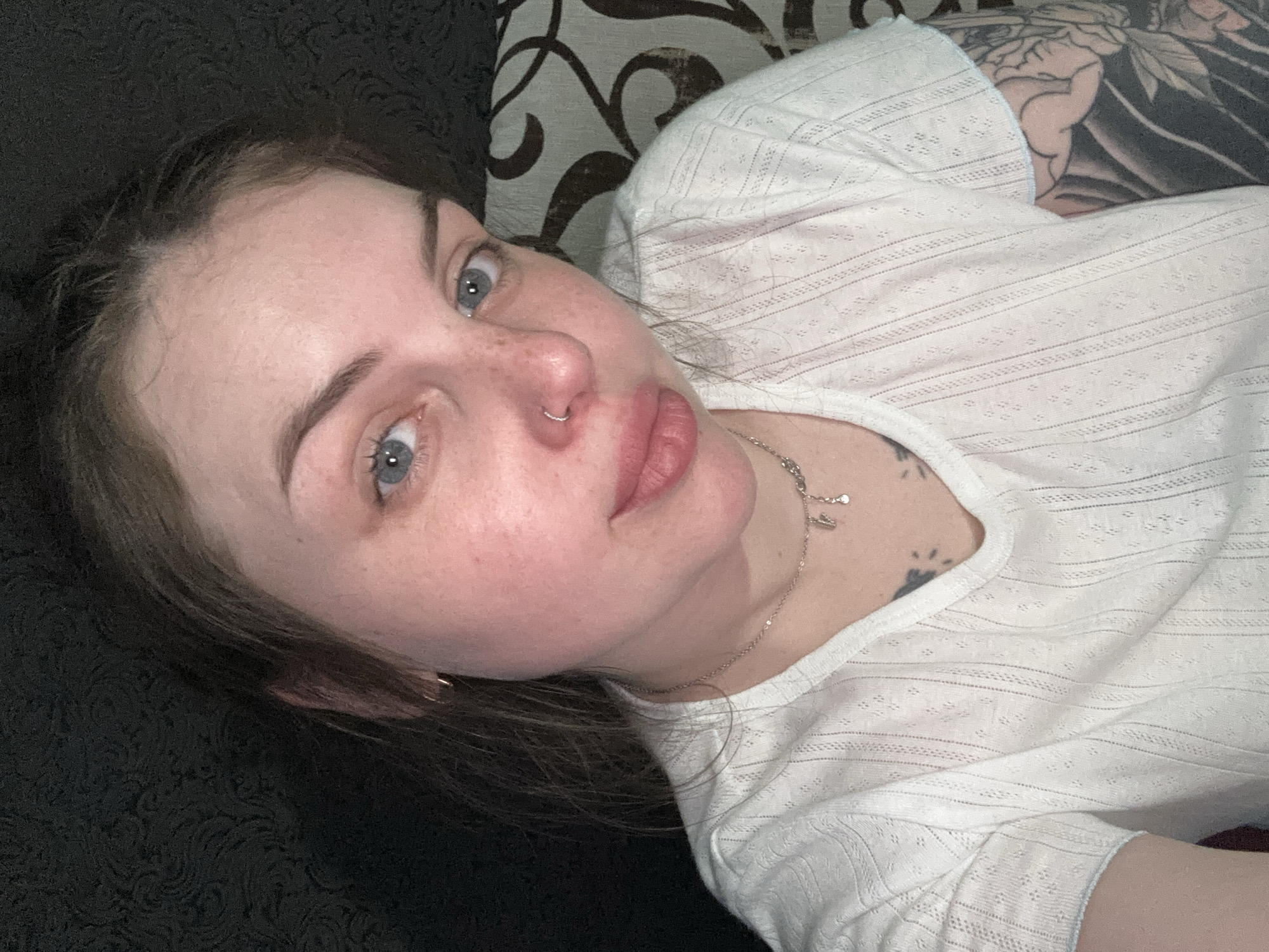 A young woman with fair skin and blue eyes is lying down, possibly on a couch or bed. She is wearing a white top and a silver necklace. She has a nose ring. There is a tattoo visible on her upper arm. The background consists of patterned fabric. The location is not evident from the image.