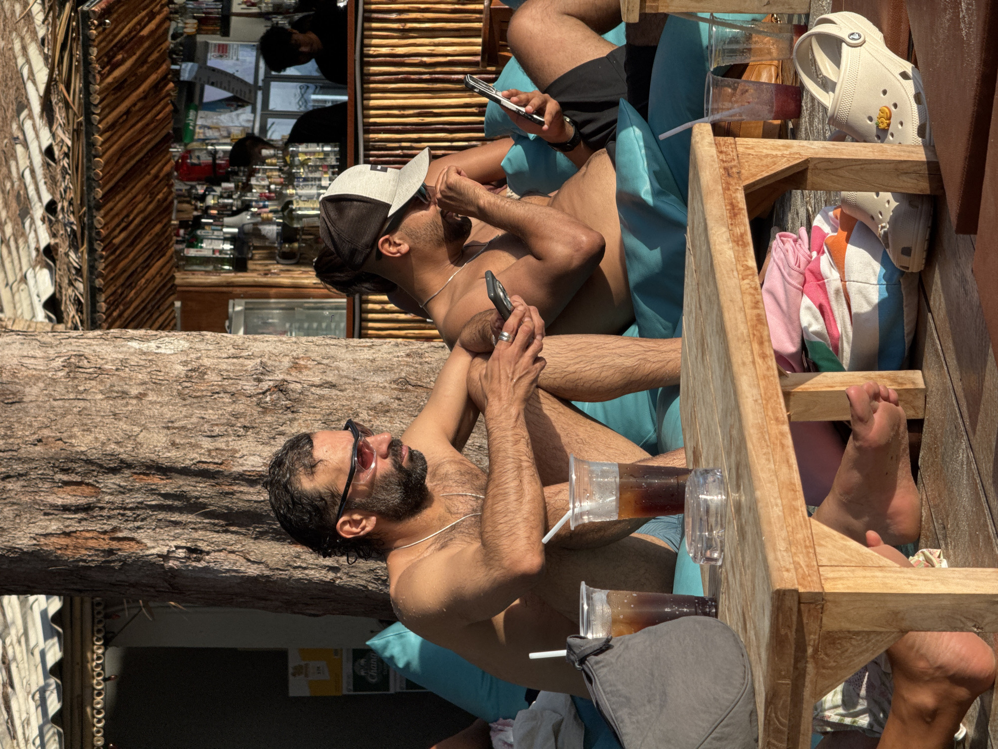 The media file depicts two individuals lounging outdoors, likely in a tropical setting given the presence of palm trees and the mention of Koh Phangan, Thailand. The scene appears to be a relaxed social gathering at a beach bar or restaurant.
The first individual, wearing sunglasses and a baseball cap, is lying down, holding a smartphone. The second individual is also reclining, with a phone in hand, and appears to be engaged with it. Both individuals are shirtless, suggesting warm weather and a casual atmosphere. Their attire, including shorts and a baseball cap, further reinforces the relaxed, beach-appropriate vibe.
Several objects are visible, including drinks in plastic cups with straws, indicating that the individuals are enjoying beverages. A pair of beige Crocs is also visible, resting on a wooden structure. Colorful folded clothing items, possibly towels or beachwear, are tucked away in a wooden shelf.
The lighting suggests it is daytime, with bright sunlight casting shadows. The overall mood conveyed is one of leisure and relaxation, with the individuals engrossed in their phones and the tranquil surroundings. The wooden elements of the furniture and the rustic structure in the background contribute to a natural, possibly bohemian, aesthetic. No visible text is discernable within the media.