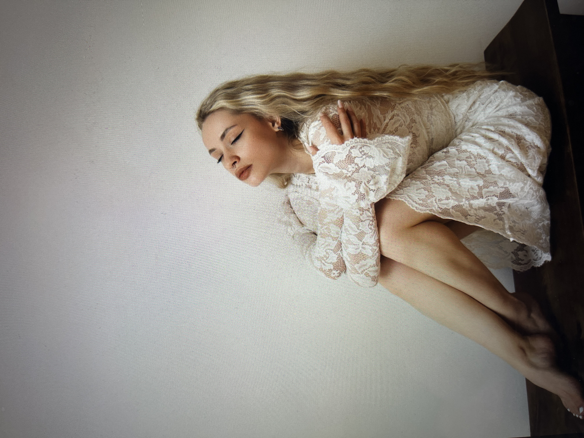 A young woman with blonde wavy hair, closed eyes, and a serene expression sits with her legs crossed on a dark wooden surface against a plain white wall. She is wearing a white lace dress with long, voluminous sleeves. Her arms are wrapped around herself, and her head is tilted back. Her makeup includes dark eyeliner and lip color. A small silver hoop earring is visible in her right ear. The lighting is soft, creating a gentle ambiance.