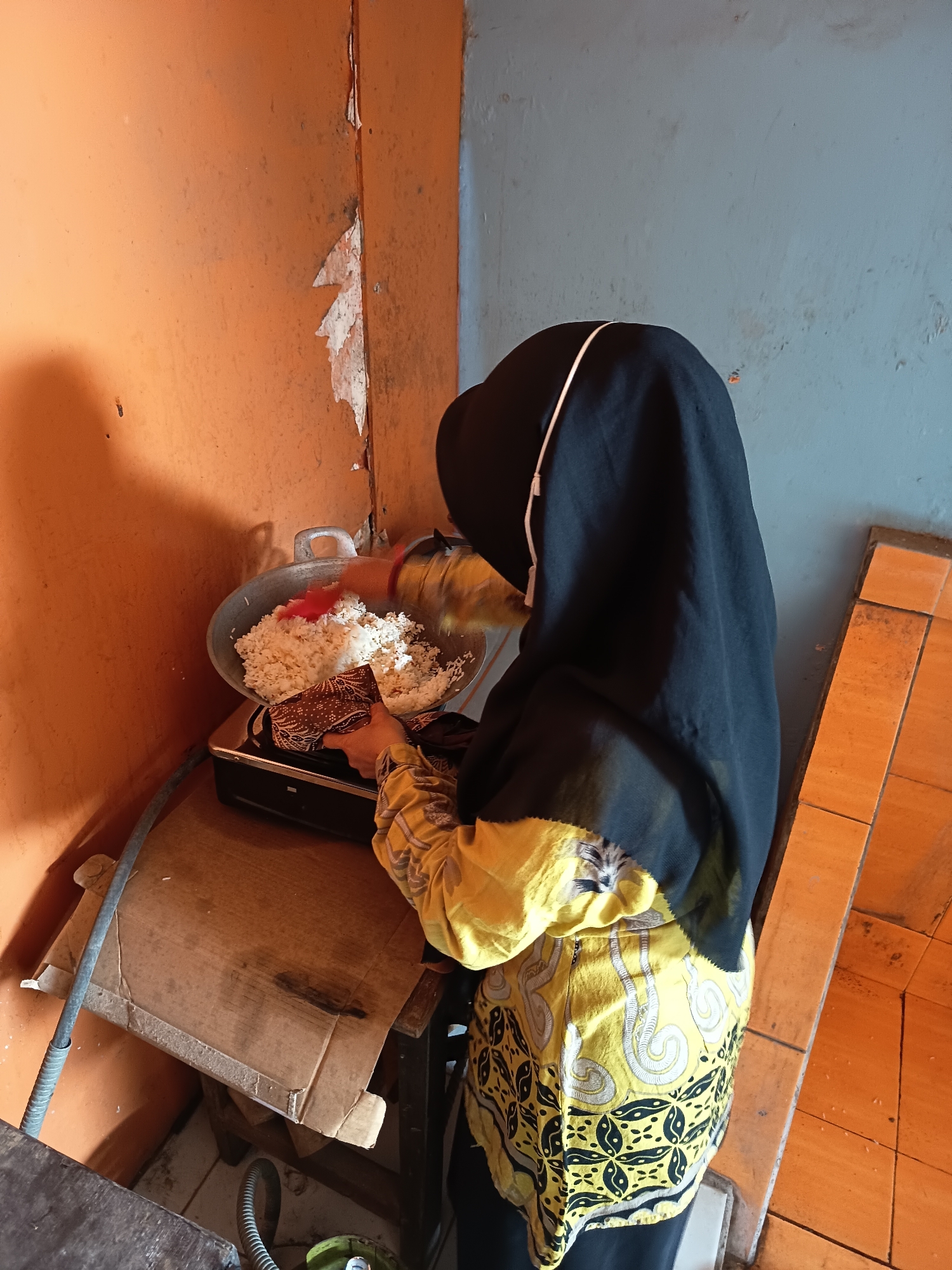 In a dimly lit kitchen area within a modest dwelling in Widoro, Indonesia, a person wearing a black hijab and a yellow batik-patterned shirt is actively engaged in cooking. They are standing over a small gas stove, where a large metal wok filled with cooked rice is placed. The person is using a red utensil to stir or mix the rice. To the left of the stove, a portion of an orange-painted wall with peeling paint is visible. The wall to the right appears to be a muted blue or gray, also showing signs of wear. The floor is covered with orange tiles. The overall environment suggests a functional but unkempt domestic space.