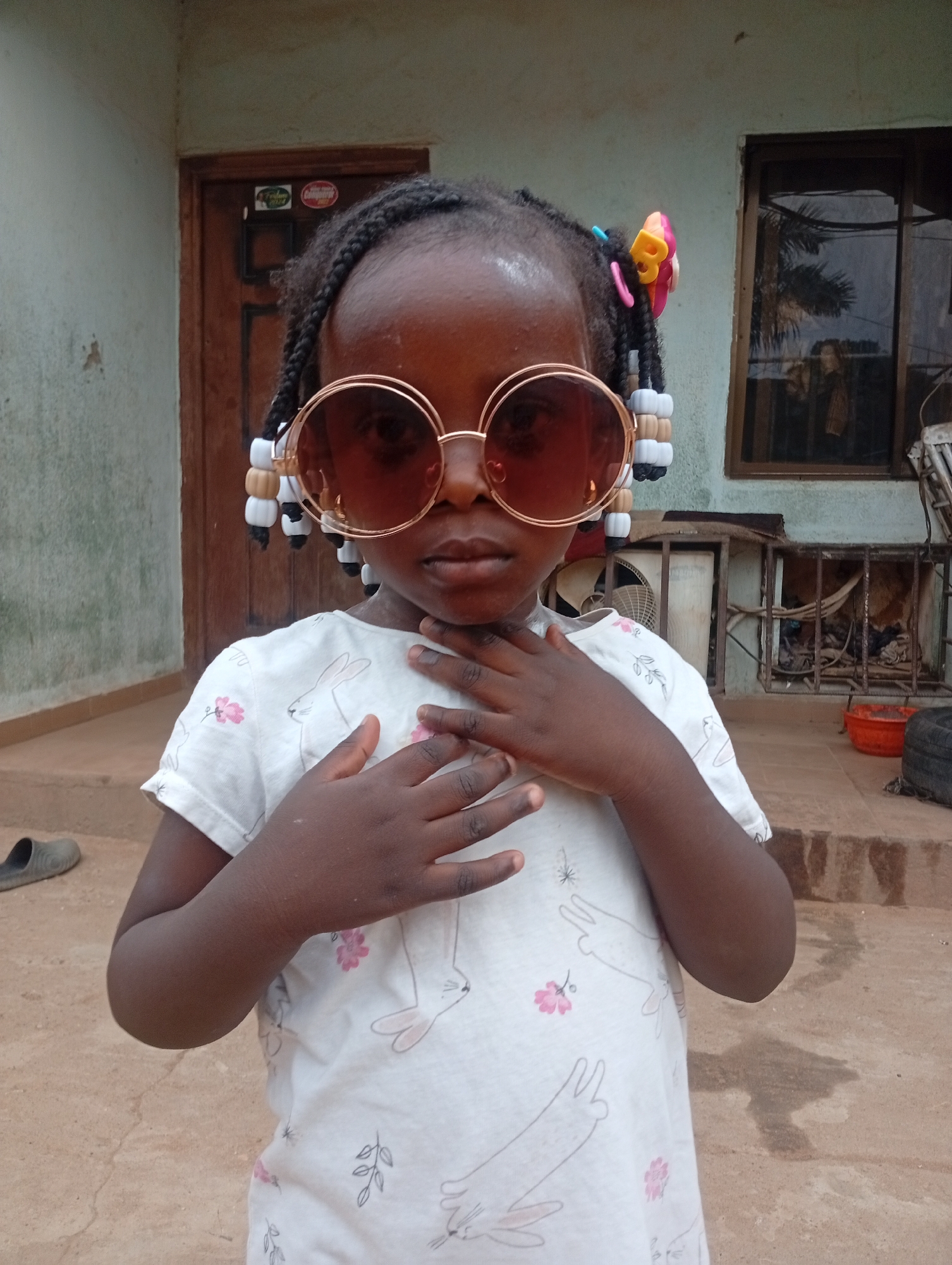 A young girl stands outside, wearing oversized round sunglasses with a rose-gold-colored frame. Her hair is styled in braids adorned with white, black, and tan beads, as well as a multi-colored hair clip. She is wearing a white t-shirt patterned with outlines of rabbits and pink flowers, and her hands are gently clasped at her chest.
Behind her, a building with a wooden door and a window can be seen. A fan and other items are also visible in the background. The ground appears to be a rough, reddish-brown surface. The location is Sabon Tasha, Nigeria.
