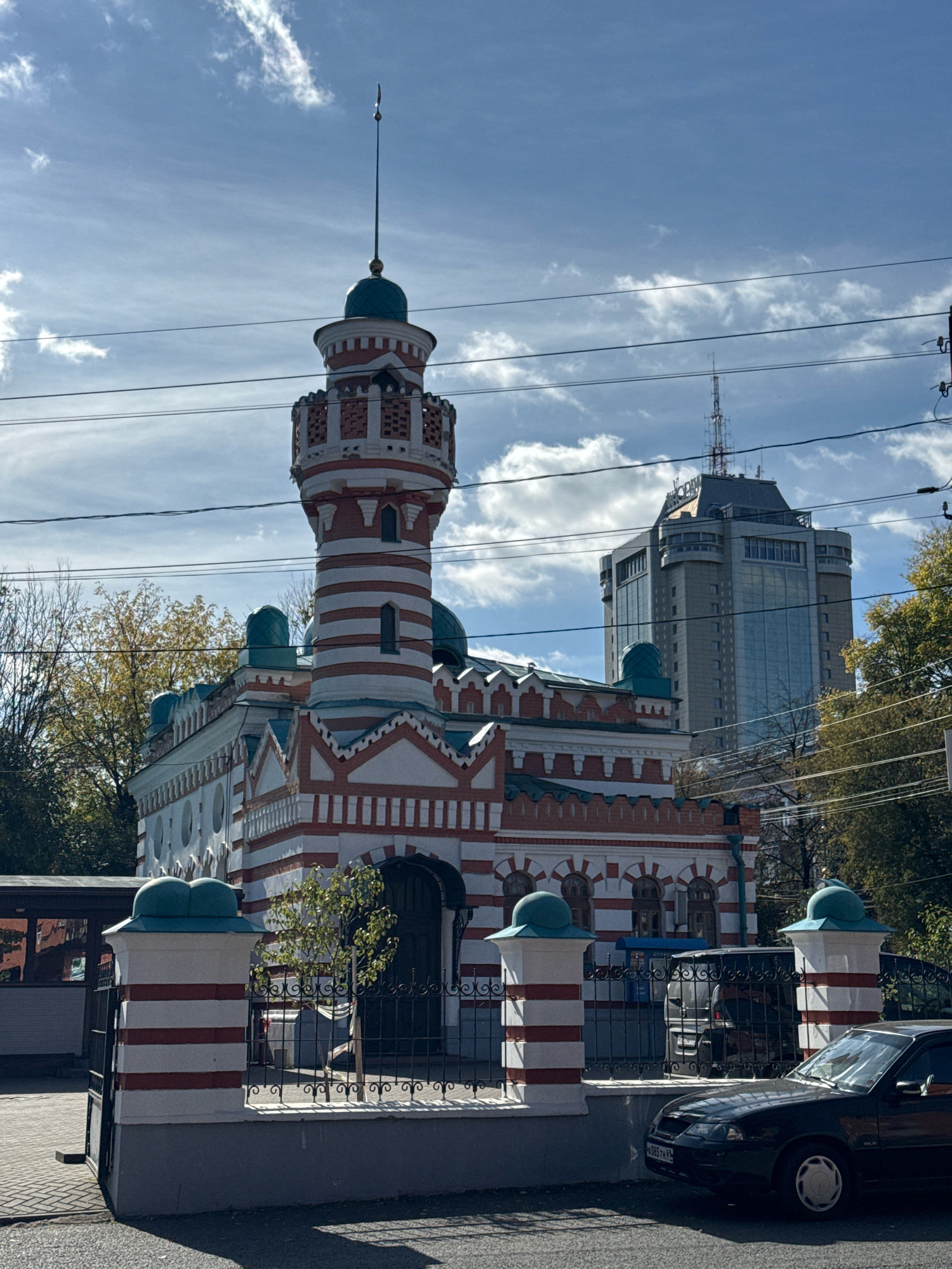 In Tver, Russia, a historical building with a distinctive red and white striped tower and green domes stands under a partly cloudy blue sky. The building features intricate white architectural details and a series of arched windows. In the foreground, a black car is parked on an asphalt road. A metal fence with red and white striped pillars encloses the property. Behind the historical structure, a modern, tall glass skyscraper with a communications tower on top is visible. Autumn foliage can be seen on trees to the right of the frame. Overhead, power lines crisscross the sky.