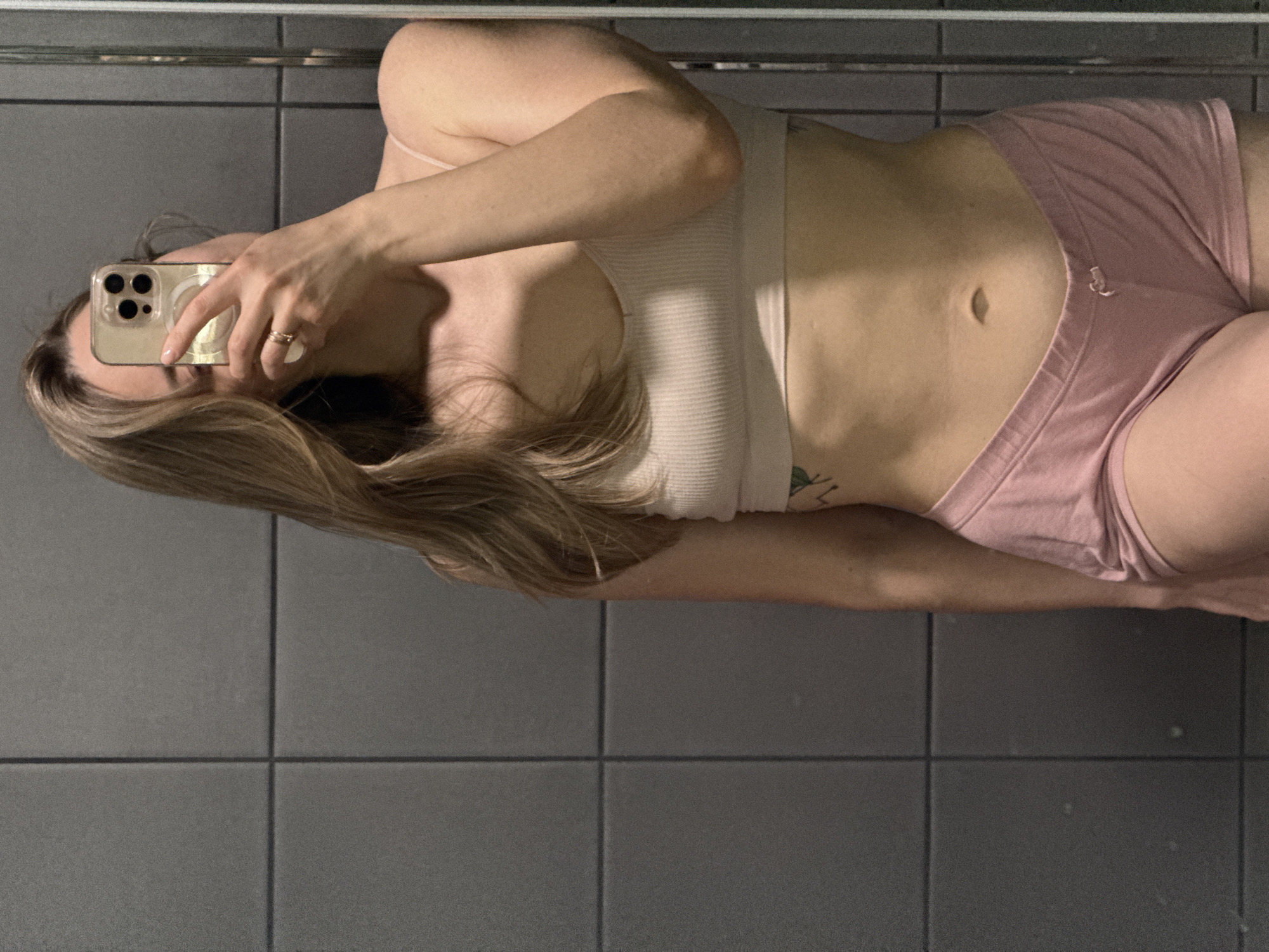 A young woman stands in a bathroom with gray tiled walls, taking a selfie in a mirror. She is wearing a white tank top and pink shorts. She holds her phone in her right hand, obscuring her face. The bathroom tiles are dark gray with thin grout lines.