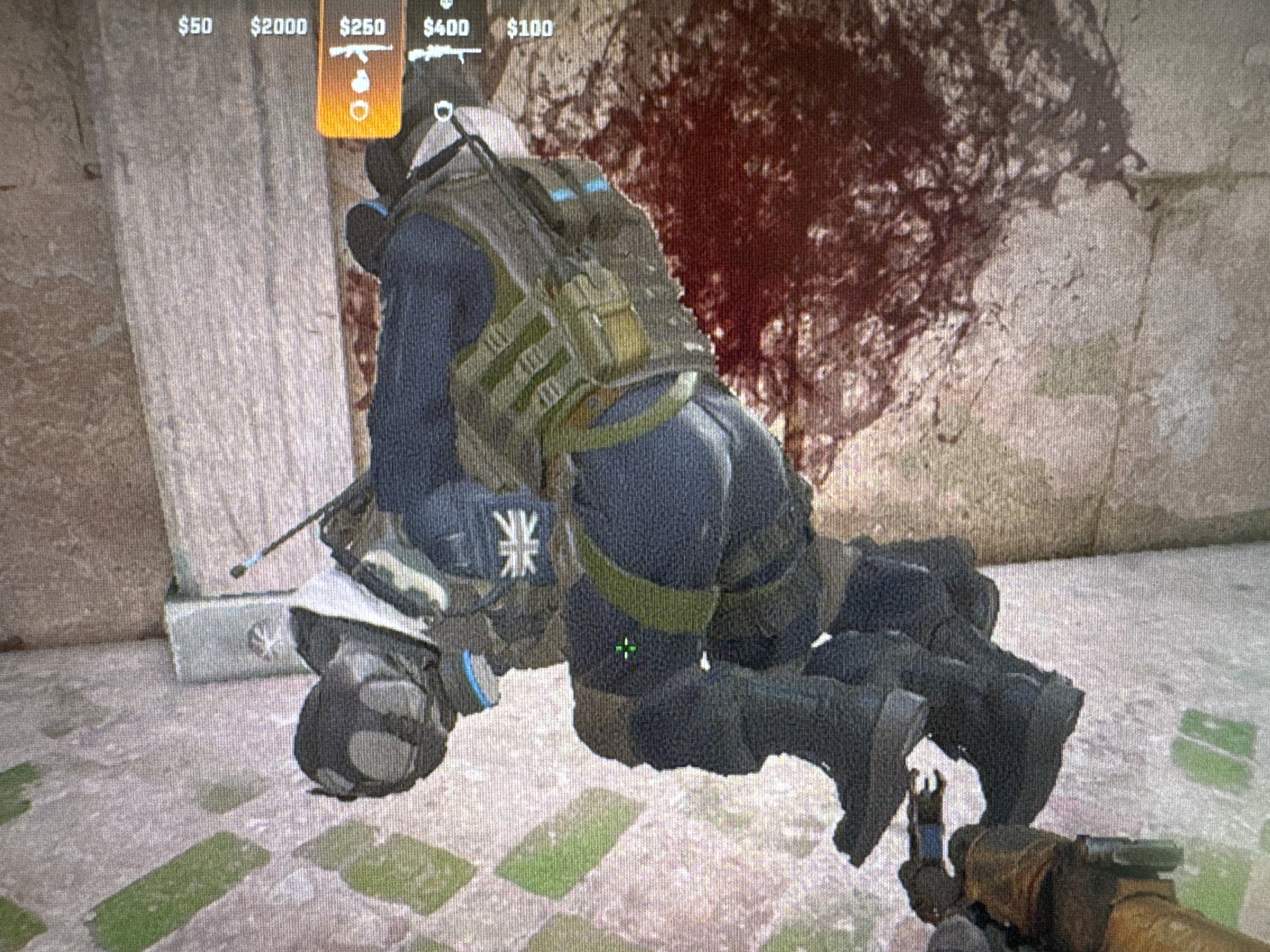 The image depicts a first-person perspective within a video game. In the foreground, the player's gloved hands are visible, holding a rifle with iron sights. The background features a scene of combat, with one character in tactical gear kneeling over another character who appears to be incapacitated or deceased. The kneeling character is wearing dark blue pants, a tactical vest with multiple pouches, and combat boots. A British flag emblem is visible on the kneeling character's left arm. The environment appears to be an urban setting with stone walls and a bloodstain on the wall behind the characters. A heads-up display (HUD) shows in-game currency and weapon information, indicating the location is within a virtual environment.