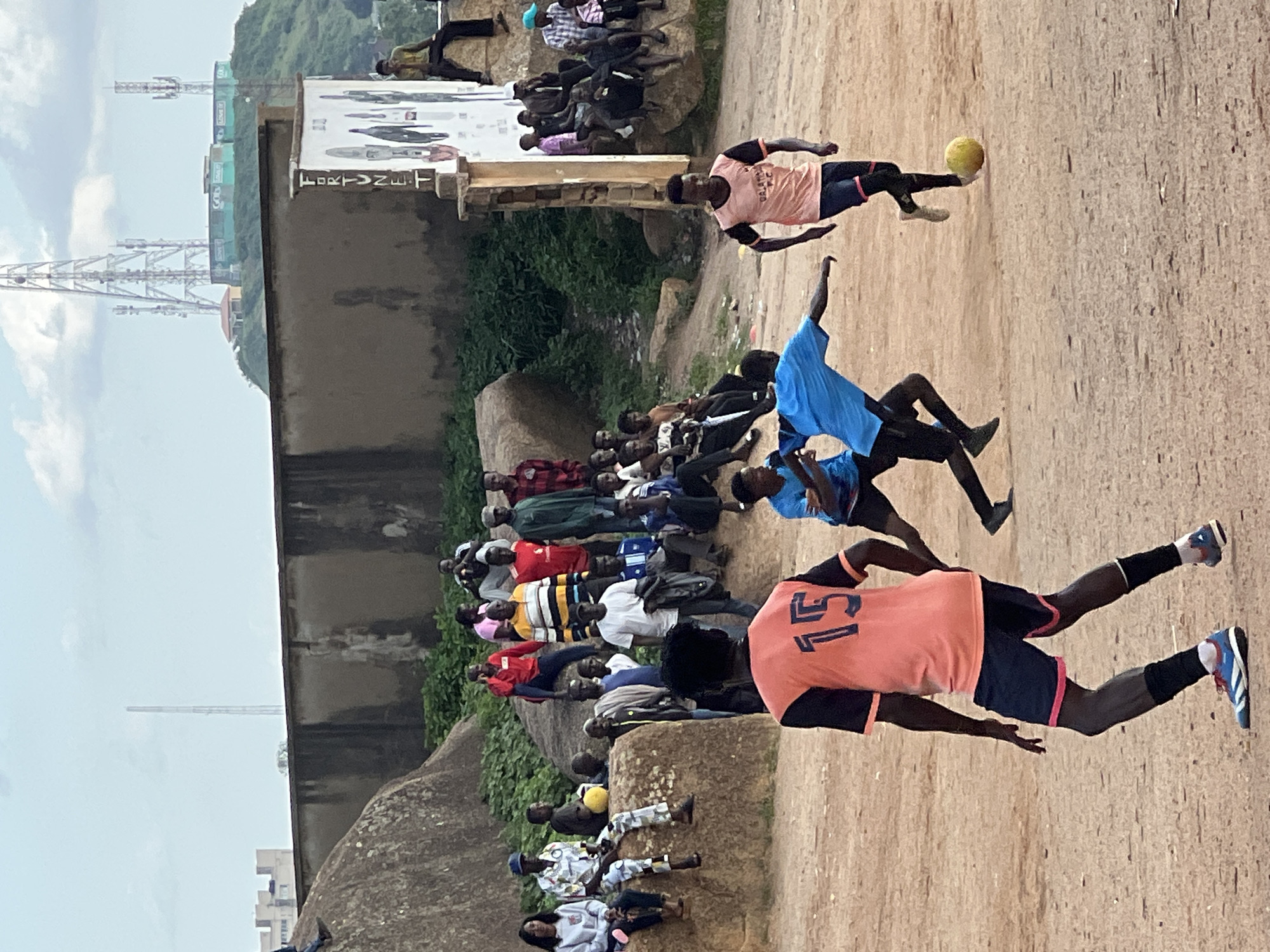 This media file depicts a group of people on a dusty, outdoor field in what appears to be an urban or semi-urban setting in Jos, Nigeria. The main subjects are individuals playing and watching a soccer (football) game. Two young men are actively playing, with one in a light pink jersey with the number "15" on the back and dark shorts, kicking a yellow soccer ball. Another player, wearing a blue jersey and dark shorts, is also in motion.
A crowd of spectators is seated on a rocky embankment and along the edge of the field, observing the game. They are dressed in casual attire, with a variety of colors and patterns visible. The background features a large concrete structure, possibly a bridge or elevated road, with several tall communication towers in the distance, suggesting a developed area. Lush green vegetation grows on the rocky terrain. The ground is covered in light brown dirt, typical of many outdoor sports fields in the region. The sky is overcast with some visible clouds, suggesting a daytime scene, though the specific time of day is not discernible. There is no strong indication of weather conditions beyond the overcast sky.
On the concrete structure, there is a large white sign with some faded text and images. The text "FORTUNE" is clearly visible in large, dark lettering on the sign. The overall atmosphere appears to be one of casual community engagement, with the focus on the sporting activity.