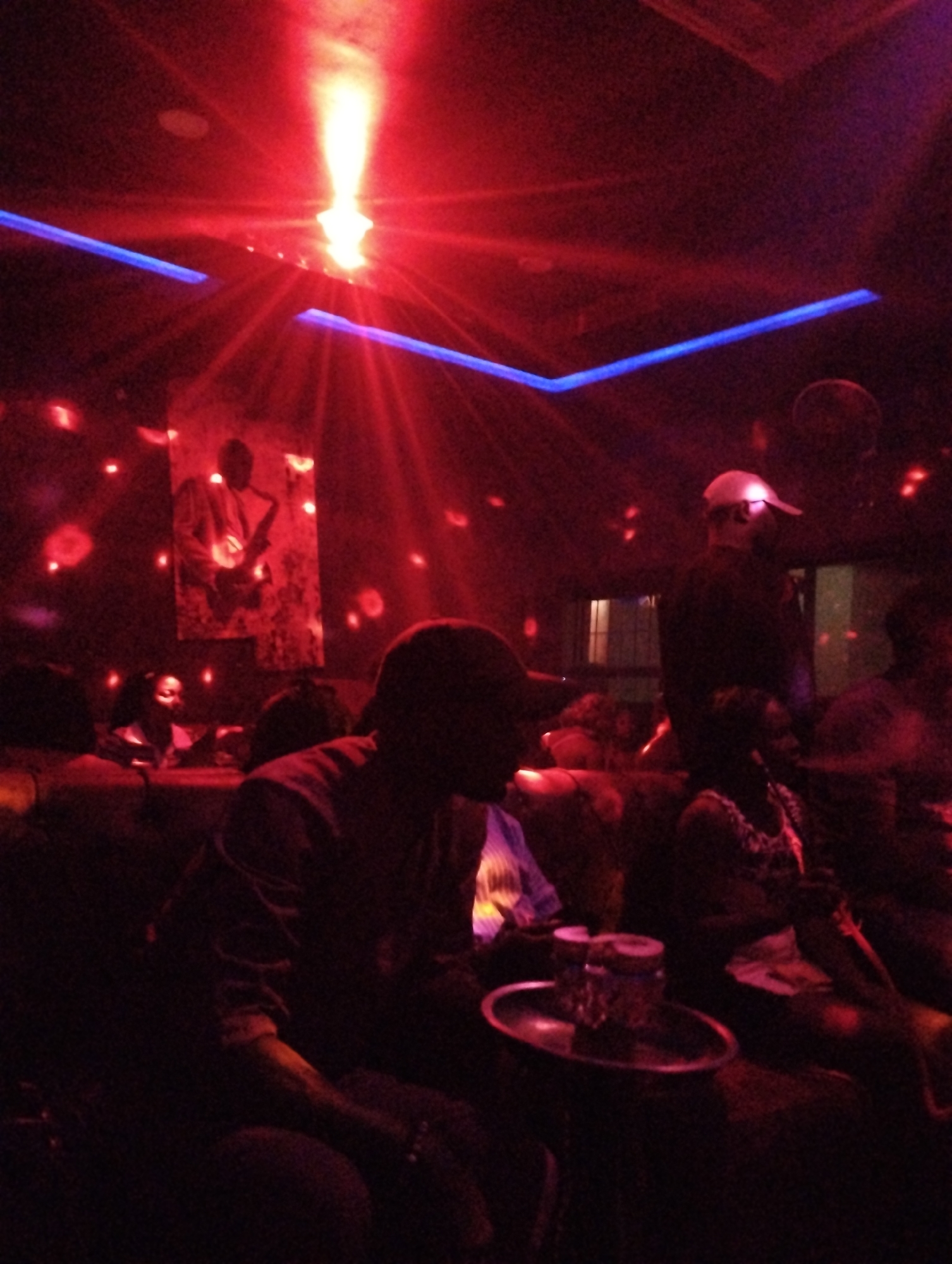 The image shows a dimly lit interior, potentially a club or lounge in Gombe, Nigeria. Red and blue lighting fixtures are visible on the ceiling. A poster or artwork featuring a musician with a saxophone is on the wall. Several people are present, seated and partially visible in the darkness. In the foreground, a person sits with a tray containing drinks. The scene is crowded and appears to be a social setting.