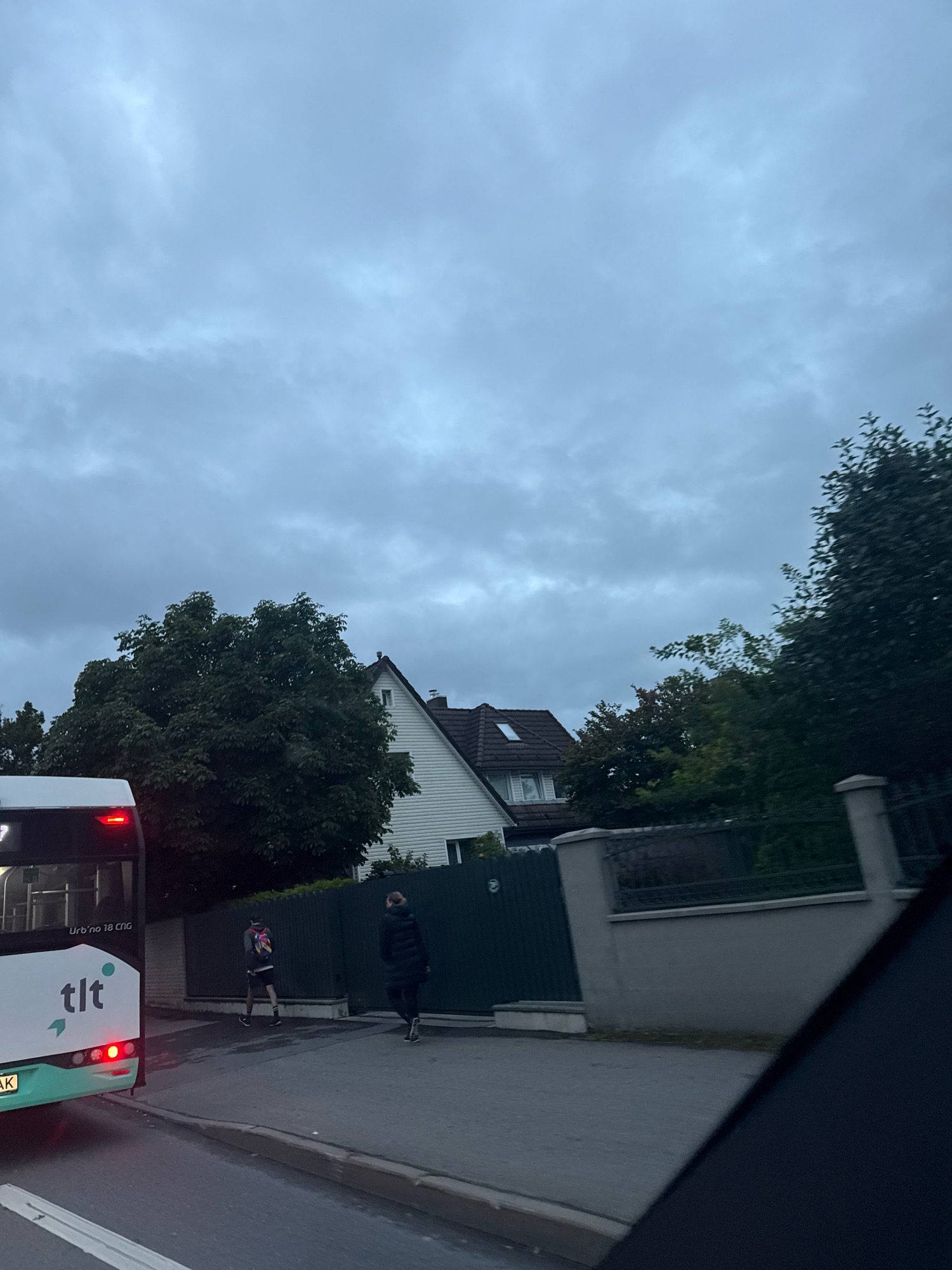 The image captures a street scene in Tallinn, Estonia, under an overcast sky. In the foreground, a white and teal bus with the "tlt" logo is partially visible on the left, its brake lights illuminated. Two individuals are walking on the sidewalk. The person on the left, a male, wears shorts, a backpack, and appears to be walking away from the bus. The person on the right, a female, is wearing a dark hooded jacket and pants, also walking away from the bus. Behind them, a dark green fence runs along a property line, with a grey stone wall and fence above it. A white-sided house with a dark tiled roof and a prominent dormer window is visible behind the fence and trees. Lush green trees partially obscure the house and the sky. The overall ambiance is muted due to the cloudy weather.