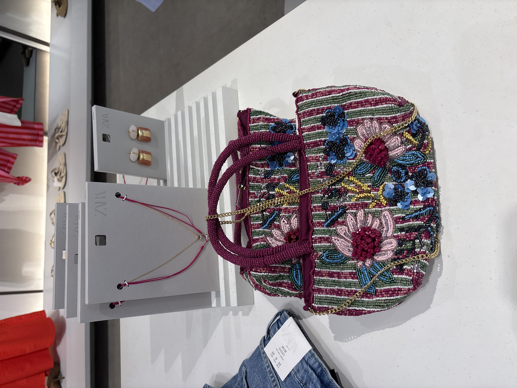 A highly detailed, close-up, top-down shot features a beaded handbag with a floral design placed on a white countertop. The handbag has a magenta top handle and a gold chain strap, with a vibrant pattern of pink, blue, and green beads forming floral motifs and vertical stripes. Beside the handbag are two grey display cards, one with the Zara logo and a delicate gold necklace, and another displaying a pair of earrings with a gold and white design. To the left of the display cards, a section of blue denim jeans with a white tag is visible. The background is blurred, showing hints of clothing racks and other retail items, suggesting a store environment. The lighting is bright, casting shadows on the countertop, with no people present in the frame. The scene is set within a retail store in Orihuela, Spain.
