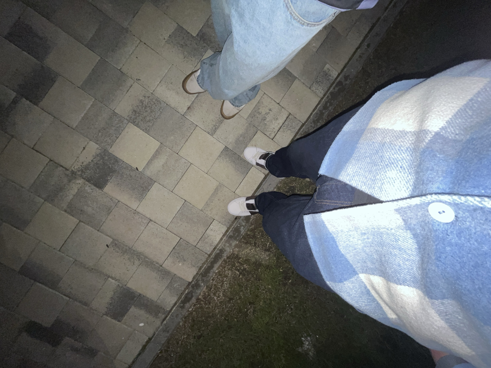 An overhead, low-light image depicts the lower torsos and legs of two individuals standing on a paved surface adjacent to a narrow strip of dormant grass. The person in the foreground wears dark blue denim jeans and a light blue and white plaid outerwear garment with a visible white button, paired with white sneakers featuring brown side panels and light brown soles. The second individual, positioned further back to the right, wears light blue denim jeans and light beige sneakers with brown soles. The paved surface consists of grey and beige rectangular blocks arranged in a diagonal grid pattern, showing varying degrees of wear and moisture. The scene is outdoors, indicating nighttime conditions in Unknown, Unknown.