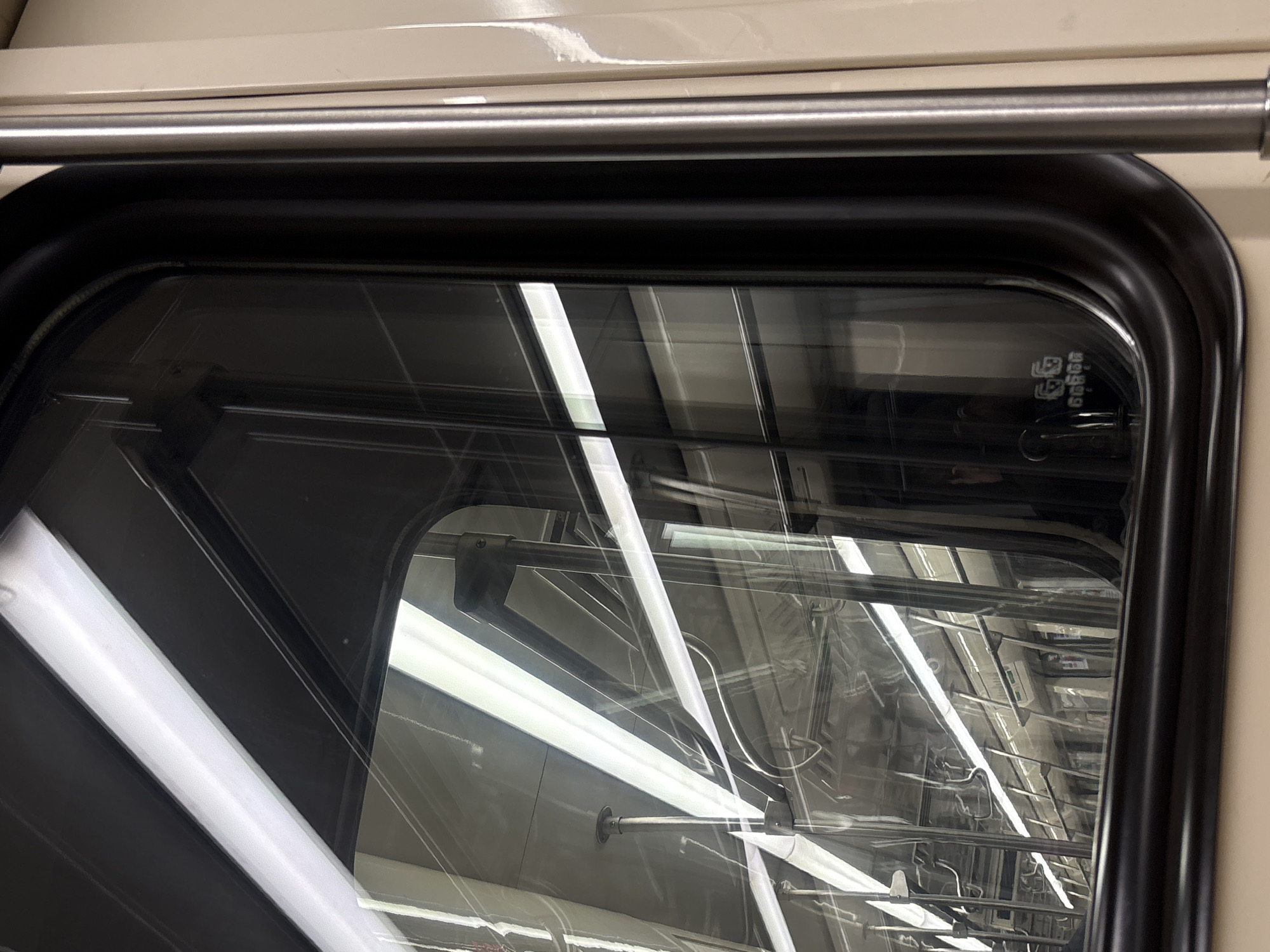 The image captures a close-up view of the interior of a metro or train car, specifically focusing on a dark-framed window or transparent panel. The most prominent feature is the heavy reflection on the glass, which almost entirely obscures any view outside and instead reveals the interior of the train itself.
Through the reflections, several long, bright fluorescent light fixtures are visible, running horizontally across the ceiling of the train car. A complex network of metal handrails, support rods, and other fixtures typically found in public transport vehicles can also be seen, suggesting an overhead luggage rack area or grab bars for standing passengers. The train's interior appears to be in shades of light beige or off-white for the ceiling and walls, with darker elements for mechanical components.
The window frame is dark, possibly black or very dark gray, with softly rounded corners. A horizontal, polished metallic bar or handrail runs just above the window, spanning the top of the frame. In the upper right corner of the transparent panel, small, faint symbols or logos are etched into the glass, possibly indicating manufacturing details or safety standards. No people are visible, nor is any specific activity taking place beyond the implied state of being inside a stationary or moving train. The lighting suggests an indoor setting, likely during any time of day, given the artificial illumination. The context provided indicates this is Saint Petersburg, Russia.