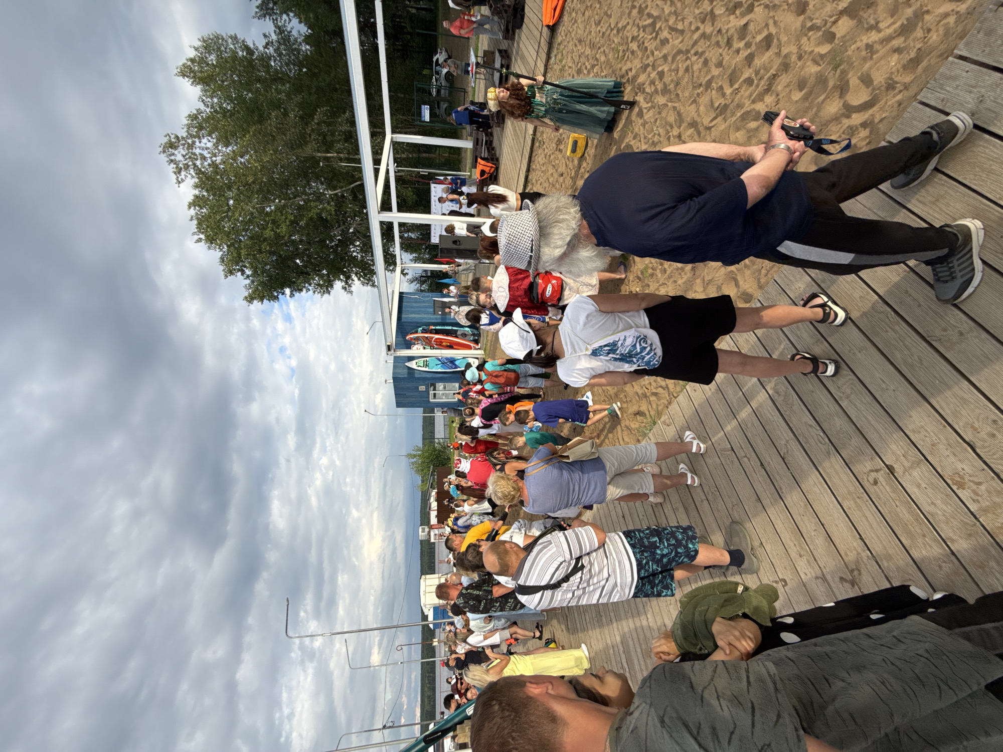 This is a media file depicting an outdoor gathering on a wooden boardwalk near a sandy area, likely a beach or waterfront. A crowd of people, of various ages and genders, are present, some standing and others seated. The scene appears to be a public event or gathering, possibly a festival or competition, as evidenced by the presence of several paddleboards and a small stage-like structure in the background.
The people are dressed in casual summer attire. Some are actively engaged, watching an event or listening to someone speak, while others are simply observing. The overall atmosphere seems relaxed and communal.
Notable details include the overcast sky, suggesting a cloudy or partly cloudy day. The presence of trees in the background and the wooden boardwalk indicate a natural or recreational setting. The time of day appears to be late afternoon or early evening, given the lighting.
While no specific text is clearly legible on any signage within the image, the context of a public gathering suggests the possibility of event-related announcements or branding. The scene is shot from a slightly elevated perspective, looking down on the crowd. The image is oriented vertically.