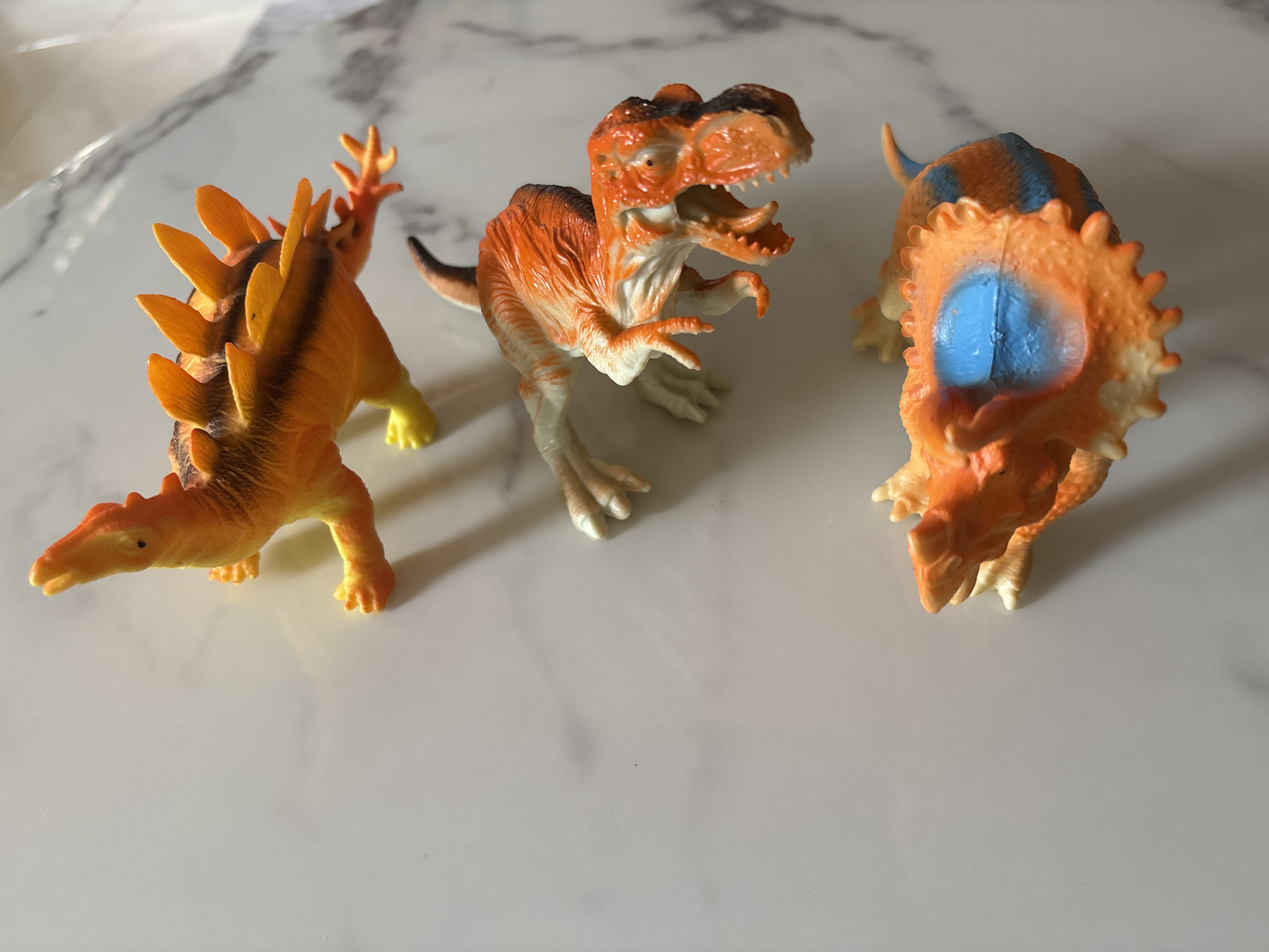 Three colorful plastic toy dinosaurs are arranged on a shiny, light-colored surface, possibly a marble or quartz countertop. The dinosaur on the left resembles a Stegosaurus, predominantly orange and yellow, with prominent spiked plates along its back and tail. In the center, facing forward with its mouth open, is an orange and white Tyrannosaurus Rex-like figure, showing detailed teeth and clawed hands. The third dinosaur on the right, viewed from the back, is an orange and white Triceratops-like creature, featuring a large frill with a distinctive bright blue patch and blue stripes along its back. The surface reflects light, suggesting an indoor setting under bright illumination, which casts subtle shadows behind the toys. No specific activity is depicted, as the dinosaurs are static objects. There is no visible text in the image. The scene is located in Abuja, Nigeria.