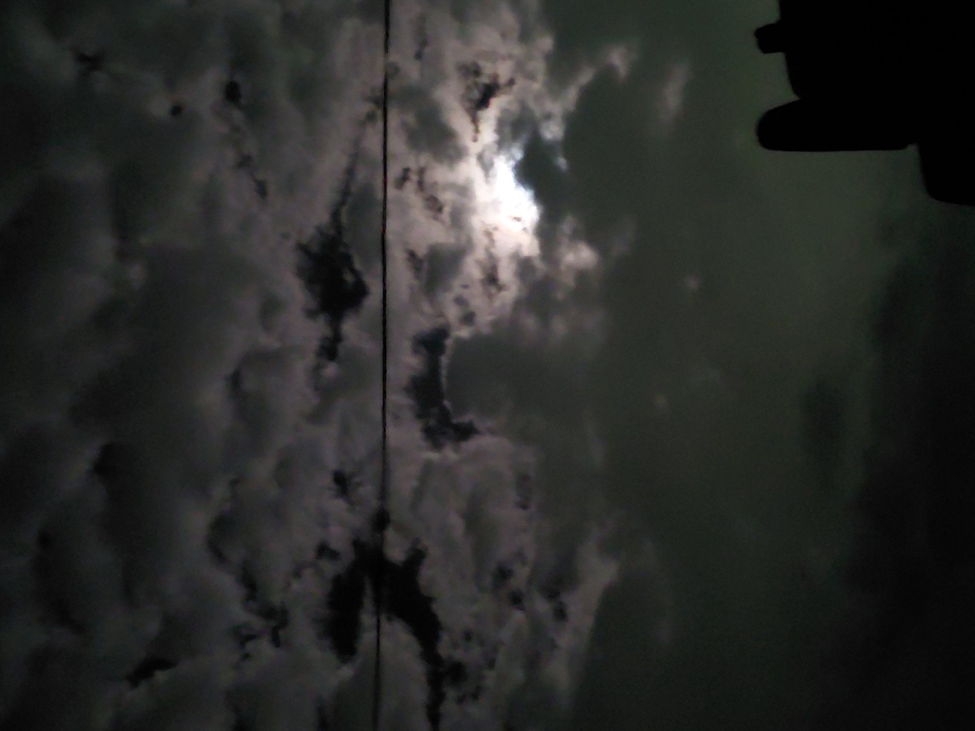 The media file captures a dramatic nighttime scene of a heavily clouded sky, illuminated by a bright, obscured light source, likely the moon, peeking through the dense cloud cover. The sky is predominantly dark grey and black, with brighter white and light grey tones around the central light source where the clouds reflect the light intensely. A thin, dark, vertical line, possibly a utility wire or cable, cuts across the mid-left side of the frame. In the upper right corner, a dark, abstract silhouette of an unknown object or structure is partially visible, suggesting it is a foreground element from the perspective of the photographer. The overall impression is one of an overcast night. No individuals or discernible text are present. The scene is set in Santa Ana, Venezuela.