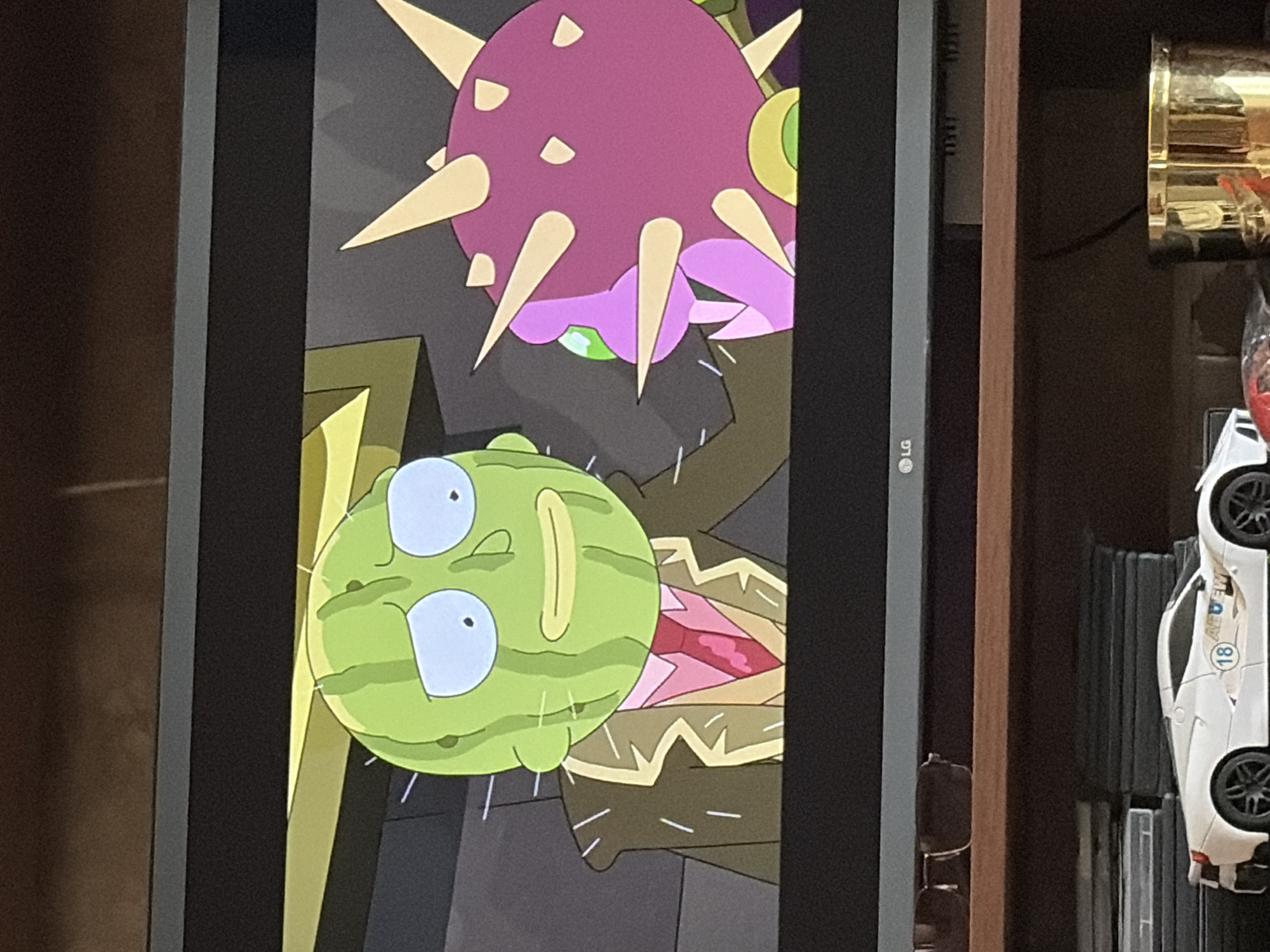 A television screen displays an animated scene from "Rick and Morty." On the left, a character with a green, watermelon-like head wearing a suit and tie looks distressed. On the right, a purple, spiky creature with yellow spikes and a green eye appears to be interacting with the watermelon-headed character. The background is a dimly lit, abstract environment with yellow and grey elements. Below the television, a shelf holds a white toy sports car and other objects.