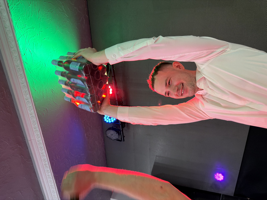 An indoor scene within a club or event venue in Votkinsk, Russia, depicts a male individual, appearing to be in his late 20s or early 30s, with short hair and a white long-sleeved shirt, smiling broadly. He stands with both arms extended straight overhead, holding a shrink-wrapped package containing twelve glass bottles, arranged in a 3x4 configuration, with light-colored labels and red caps. To the left, a blurred arm and clenched fist, belonging to a second individual, are visible and raised. The background consists of a dark wall with at least two LED stage light fixtures emitting blue and red light. The ceiling, featuring decorative molding, is illuminated by a green light source. The overall lighting is dim, accentuated by the colored stage lights. The primary subject's expression and posture indicate a celebratory action.