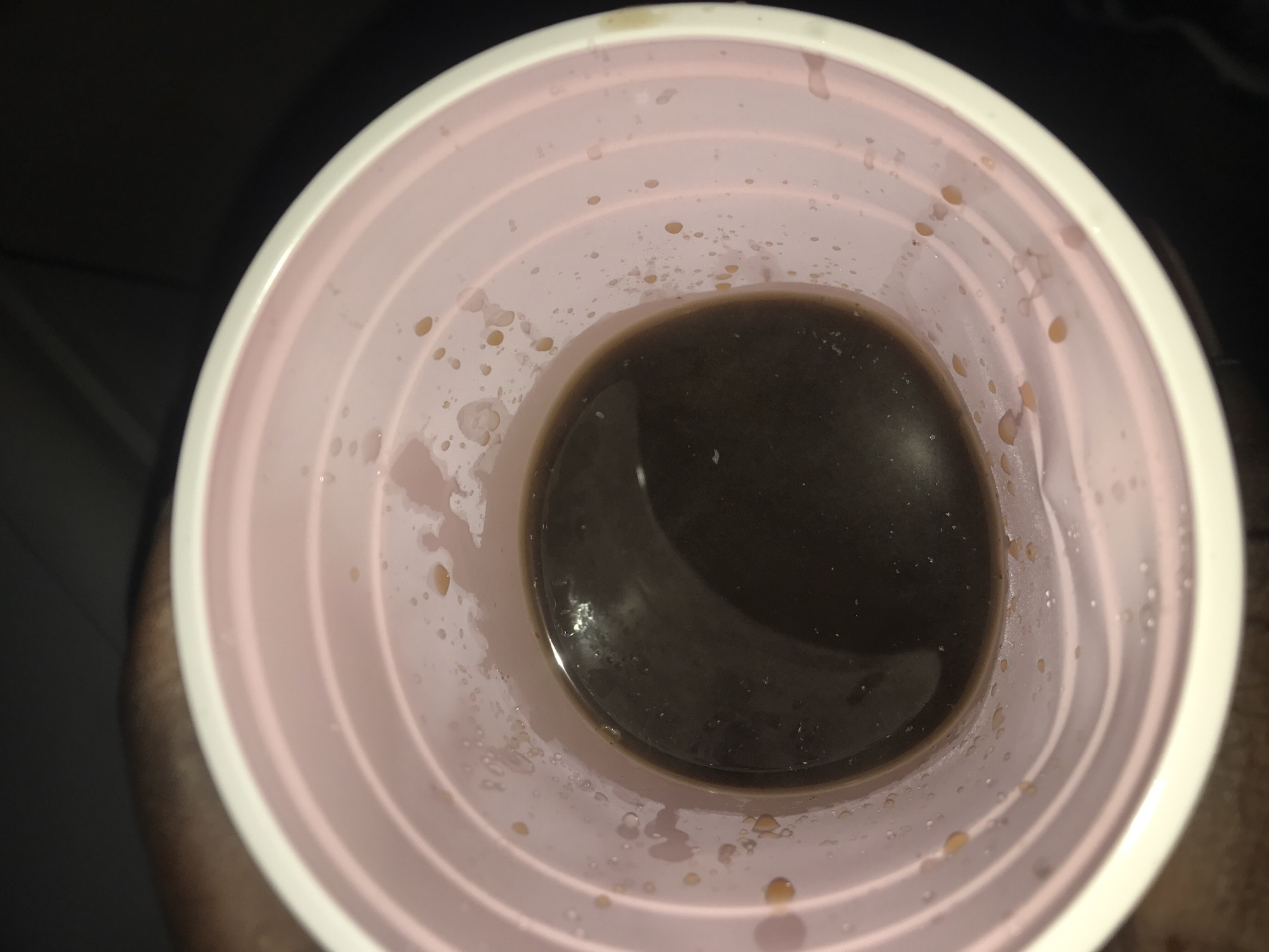 The image shows the inside of a pink plastic cup containing a dark brown liquid, likely coffee or tea. The cup has visible concentric rings on its interior surface and appears to be slightly wet, with small droplets of liquid scattered on the inner walls above the liquid level. The background is dark and out of focus, suggesting low lighting conditions. There is no discernible view of New Stadium, Nigeria, or any other specific location within the city. The image focuses solely on the contents and interior of the cup.
