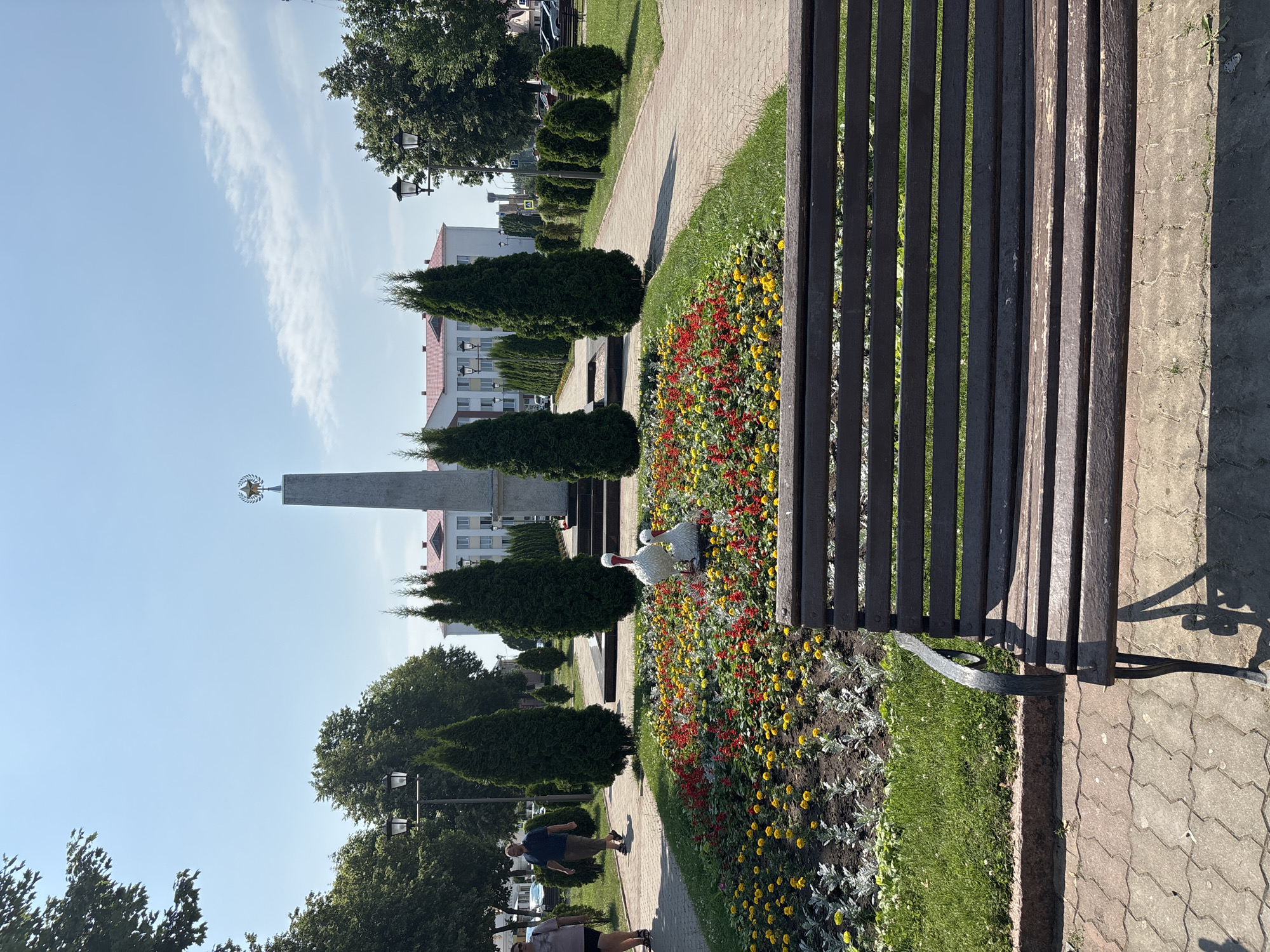 The image captures a view of a park or town square in Pastavy, Belarus, on a clear, sunny day. In the foreground, a dark wooden park bench with metal armrests sits on a paved walkway. Beyond the bench, a flowerbed bursts with red and yellow flowers, interspersed with foliage. Two white stork figurines, one larger and one smaller, are placed within the flowerbed.
A paved pathway curves to the left, where two individuals are walking. Further into the scene, a tall, obelisk-shaped monument stands prominently, topped with a golden emblem. Lined by manicured evergreen trees on either side, the monument is situated in front of a white building with a red roof. Several streetlights are visible along the pathway. The background reveals more trees and a blue sky with scattered white clouds. The overall impression is one of a well-maintained public space.