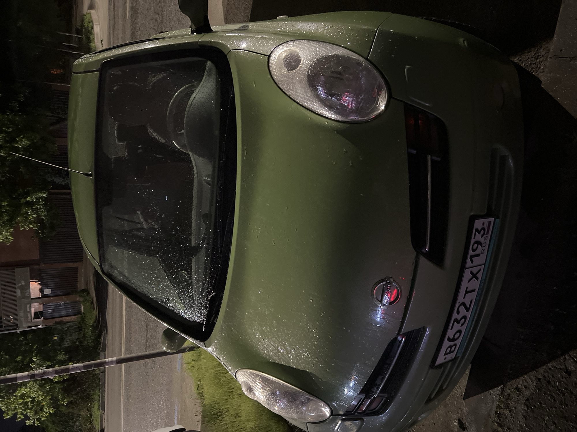A green Nissan compact car, likely a Micra (K12 generation), is depicted parked outdoors at night. The vehicle is covered in a multitude of water droplets, indicating recent rainfall or ongoing light precipitation. The scene is dimly illuminated by artificial lights, which create reflections on the wet surfaces of the car and the ground.
The photograph is taken from an elevated perspective, showcasing the front-right side of the car, including the hood, windshield, and front bumper. The car's headlights are off. In the background, dark foliage and a wet road surface are visible, characteristic of a street or parking area at night.
The car's front license plate is clearly legible, displaying the Russian registration "B632TX 193 RUS". The "193" region code indicates the vehicle is registered in Krasnodar Krai, Russia, aligning with the provided location context. The Nissan logo is also visible on the car's front grille. No people or specific activities are observable; the car appears stationary and unoccupied.