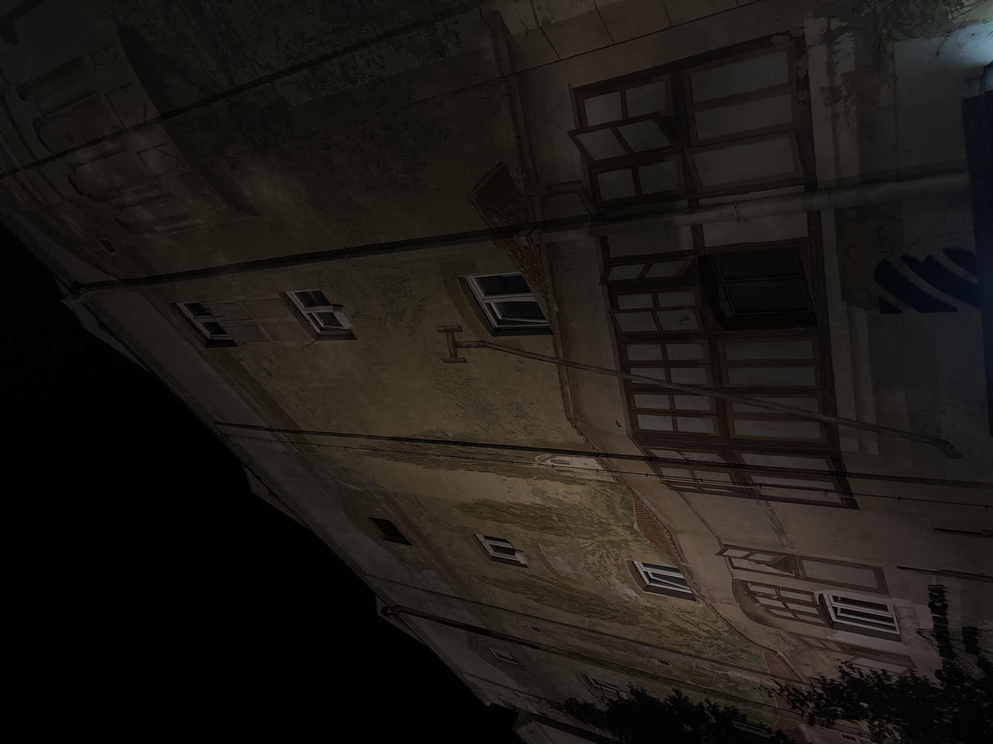 An angled nighttime shot of a building facade in Belgrade, Serbia. The building is multi-story with a weathered, textured facade. Several windows are visible, some open, some closed. There are utility lines running across the front of the building. Dark foliage is seen at the bottom right corner. The left side of the frame is mostly dark sky.