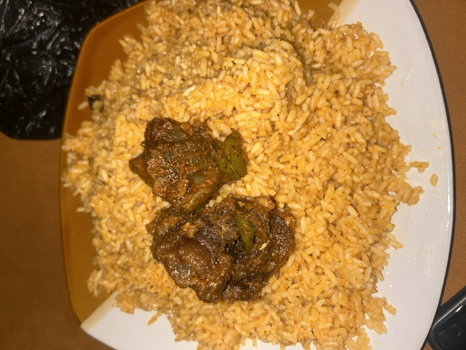 A close-up, overhead view depicts a serving of Jollof rice with two pieces of stewed meat on a square white plate with an orange-brown diagonal rim. The rice, which is reddish-orange, indicating a tomato base, occupies most of the plate. Two irregular, dark brown pieces of meat, appearing stewed or fried in a rich sauce with small green herb fragments, are positioned centrally on the rice. The plate rests on a smooth, solid brown surface, likely a table. A dark, textured object is partially visible in the upper right background, out of focus. The scene suggests an indoor setting in the city of Egbe, Nigeria.