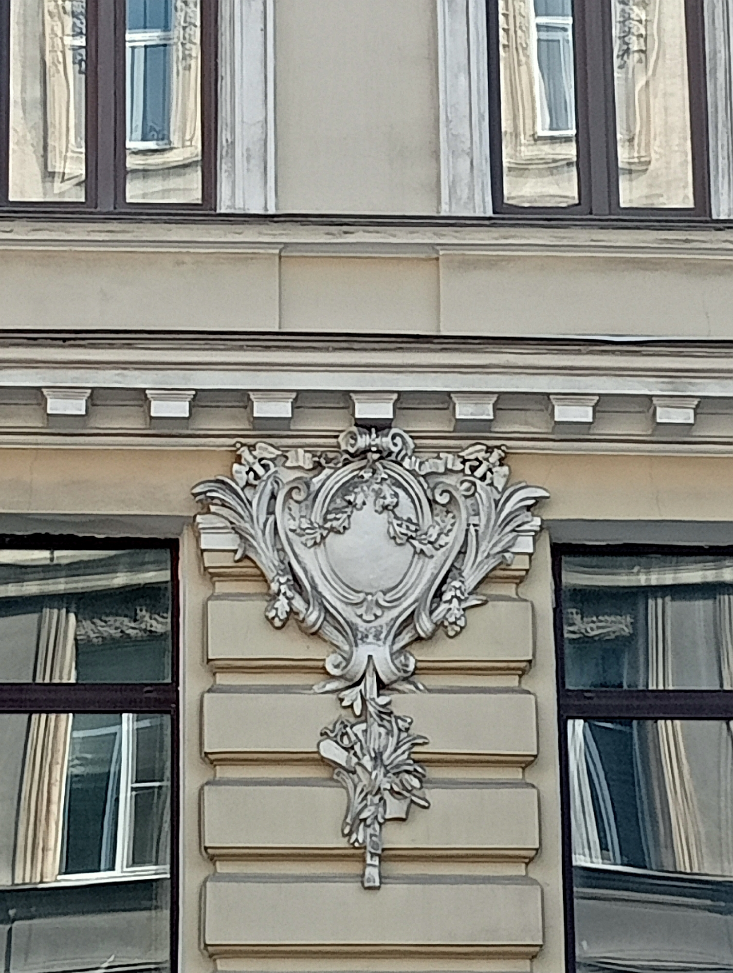 The image captures a close-up view of ornate architectural detailing on the facade of a building, likely in Saint Petersburg, Russia. The focus is on a decorative, silver-toned stucco element situated between two windows. This element is intricate, featuring a central oval shape surrounded by swirling, leaf-like motifs and floral accents. The windows on either side have dark frames and reflect the surrounding environment, showing distorted views of other windows and building elements. The facade itself is a muted beige color, with horizontal banding beneath the decorative piece. Above the decorative element, a detailed cornice runs horizontally. The overall impression is one of classical or art nouveau architectural style.