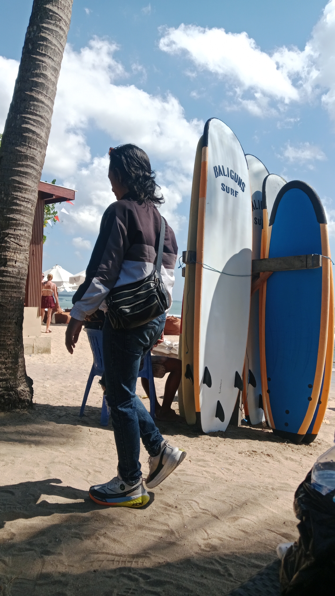 The image depicts a sunny beach scene in Kuta, Indonesia, with a focus on a person walking on the sand. A tall palm tree dominates the left side of the frame, casting a shadow. In the background, a person in a bikini and red skirt is visible near a beach umbrella and a wooden structure. Several surfboards are propped upright, leaning against each other, with "BALIGUNA SURF" branding visible on one of them. The foreground shows the sandy beach, with footprints and shadows. The sky is blue with scattered white clouds. The person in the foreground, viewed from behind and to the side, is wearing jeans, a jacket with a striped pattern, and athletic shoes. They are carrying a black fanny pack across their chest. The overall atmosphere is one of a casual beach day.