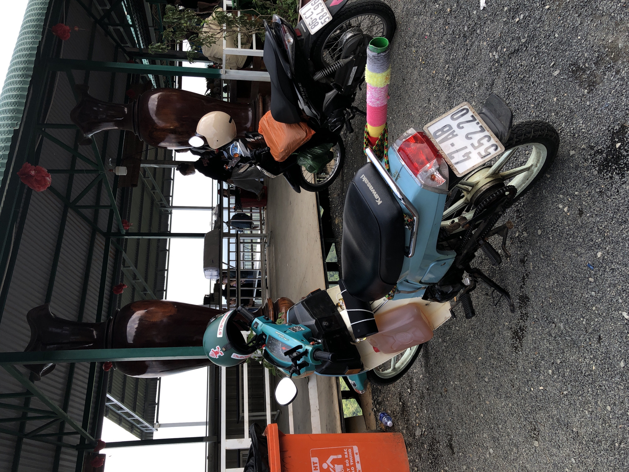 In Nam Hải, Vietnam, under a covered outdoor area supported by green metal beams, two parked motorbikes are visible on a gravel surface. The motorbike in the foreground is a light blue and cream colored scooter with a green helmet adorned with stickers placed on its handlebars. A translucent pink plastic container is attached to the side of this motorbike. A roll of colorful yarn, in shades of pink, yellow, and gray, is secured to the rear of the scooter. The motorbike in the background is black and has an orange plastic bag and a green plastic bag strapped to it. A beige helmet rests on its handlebars. Large, dark brown decorative vases are positioned behind the motorbikes. In the background, there are white tables and chairs and a railing overlooking an unseen area. An orange trash receptacle with Vietnamese text and a graphic of a person discarding waste is situated to the left of the foreground motorbike.