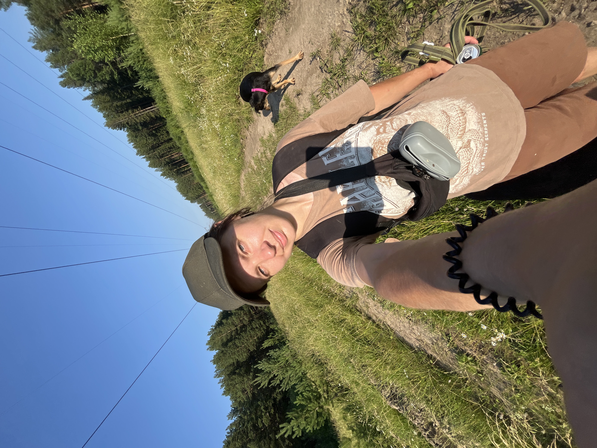 A young woman with dark hair and a green baseball cap is taking a selfie on a dirt path in a grassy field. She is wearing a light brown t-shirt, brown shorts, and a black backpack with a gray pouch attached. A small black dog with a pink collar is walking on the path behind her. In the background, there is a line of tall pine trees and a clear blue sky with overhead power lines. The sun is shining, casting shadows on the ground. The location is identified as the outskirts of Nizhny Tagil, Russia, specifically an area near power lines in a natural, undeveloped setting.
