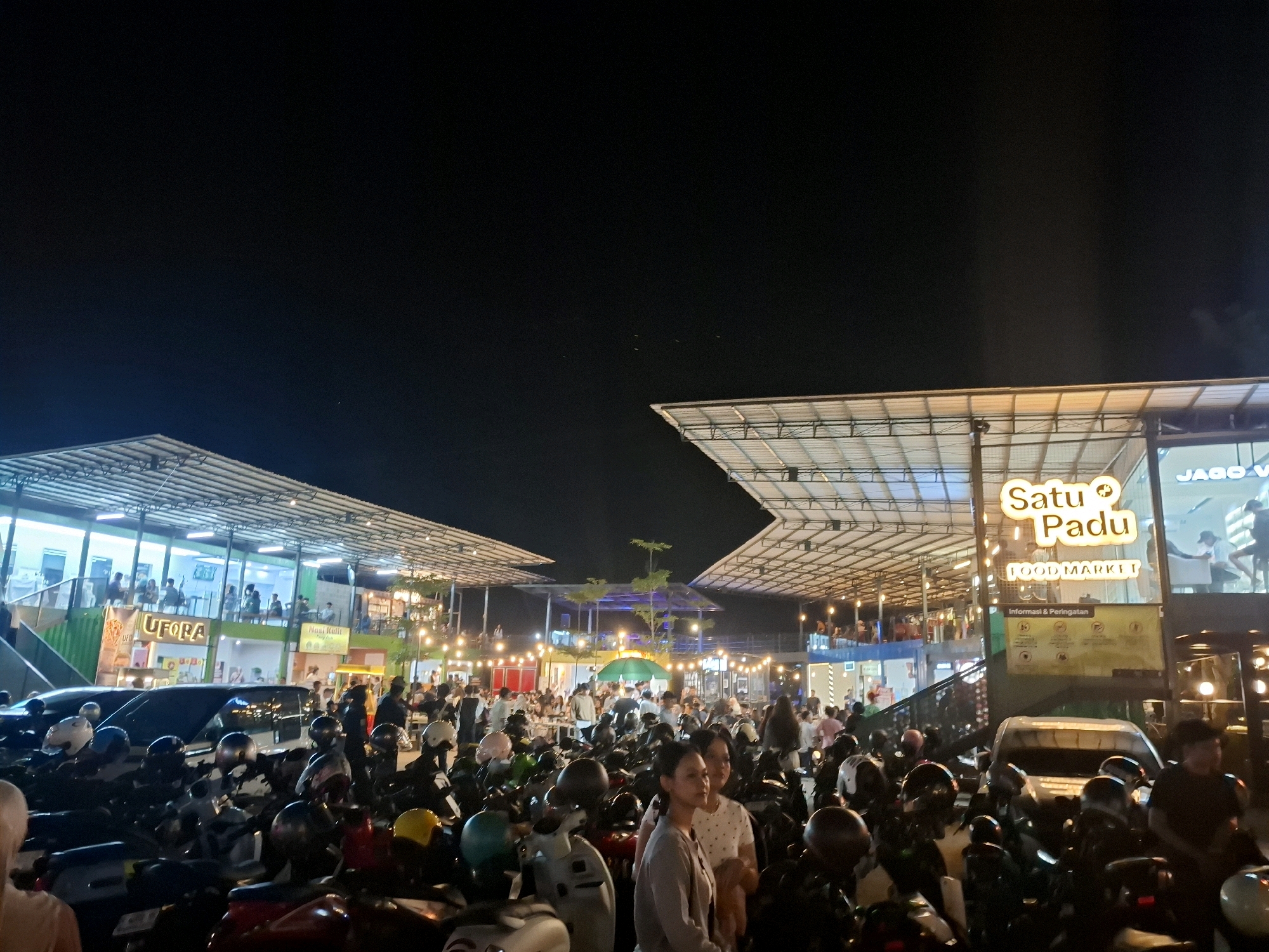 The image shows a crowded outdoor food market at night in Cianjur, Indonesia, with a large number of motorcycles parked in the foreground. The market features multiple food stalls and seating areas under covered structures with bright lighting. Signs indicate "Satu Padu FOOD MARKET" and other vendors. People are seen standing, walking, and dining. The sky is dark, suggesting it is nighttime.