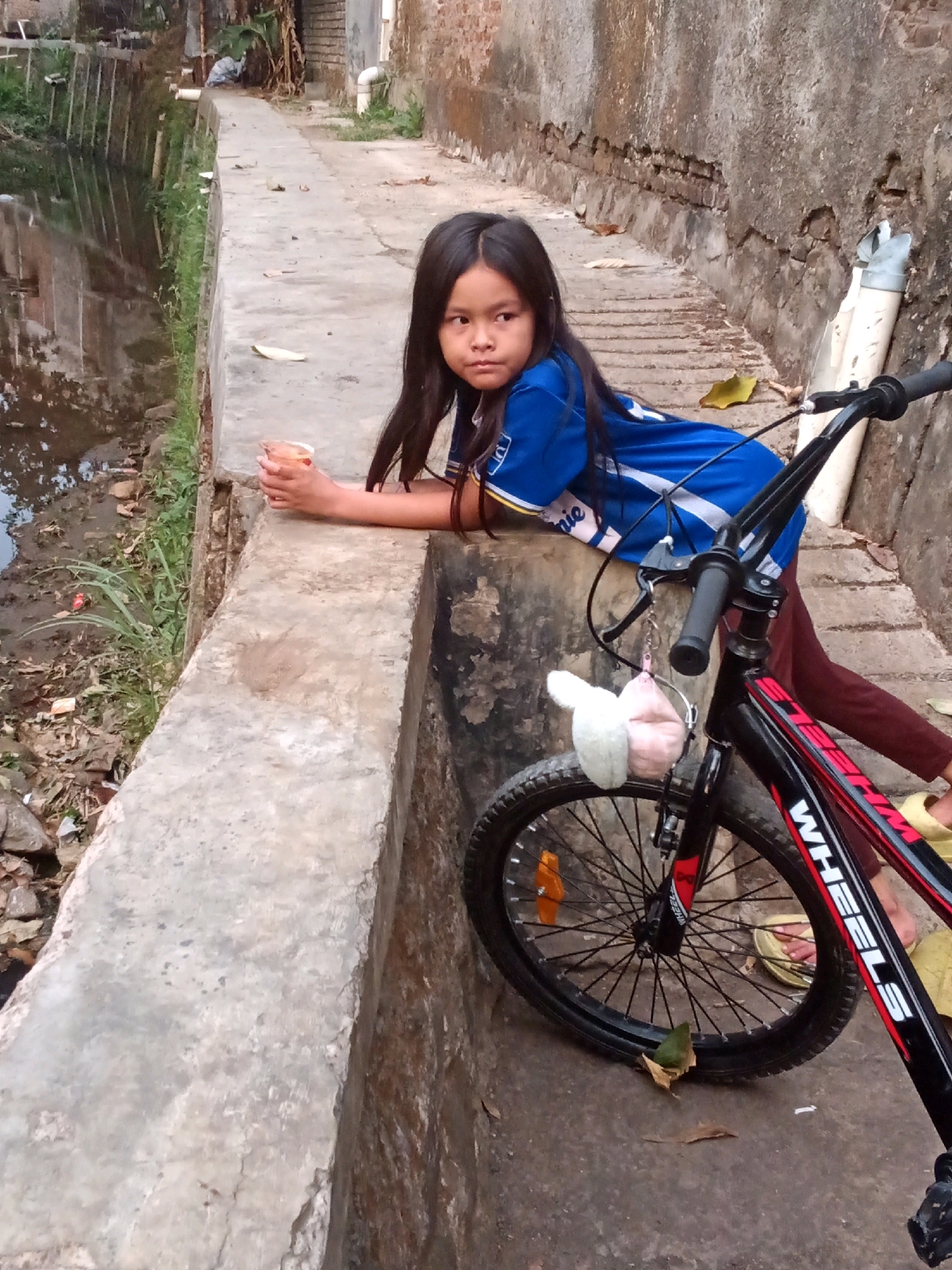 A young Indonesian girl with long, dark hair is leaning on a concrete wall next to her bicycle. She is wearing a blue t-shirt with white and yellow accents. In her right hand, she is holding a clear plastic cup with a light-colored beverage. The bicycle is black and red with "WHEELS" written on the frame. It has a bell and two stuffed toys hanging from the handlebars.
The environment appears to be an urban setting. To the left of the girl, there is a narrow body of murky water with some vegetation growing along its edge. The concrete wall she is leaning on is worn and discolored. Behind her is a brick wall with visible damage and a white pipe. The ground is paved with concrete. Fallen leaves are scattered around the bicycle. The scene suggests a casual, everyday moment in a somewhat neglected urban area. Given the details, the location is identified as a canal or drainage ditch adjacent to a residential or service area in Bandung City, Indonesia.