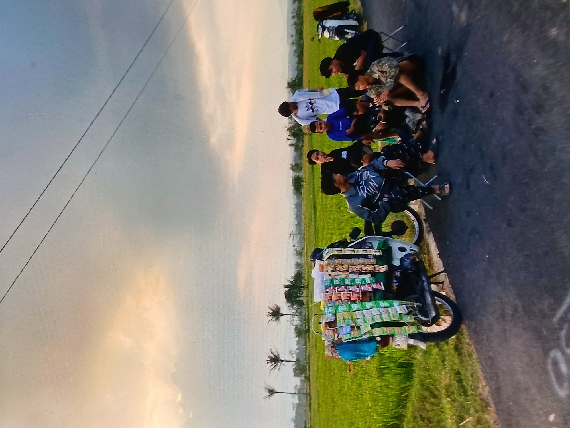A group of young Indonesian men are gathered on the side of a road next to a rice field in Setia Bakti, Indonesia, near sunset. A motorcycle, customized with a rack displaying various packaged snacks and drinks, is parked on the left side of the frame. The motorcycle is white with a black fender and exhaust. Several of the young men are seated on the asphalt, while others stand. One young man, wearing a white t-shirt, stands and appears to be talking to the others. Another young man, in a patterned jacket, is seated on a foldable chair. A white motorcycle is parked on the right side of the road, with a brown jacket draped over it. The background features a vibrant green rice field stretching to the horizon, with a few palm trees visible. The sky is overcast with dramatic clouds, tinged with the warm hues of the setting sun. The overall scene depicts a casual gathering of friends in a rural Indonesian setting.