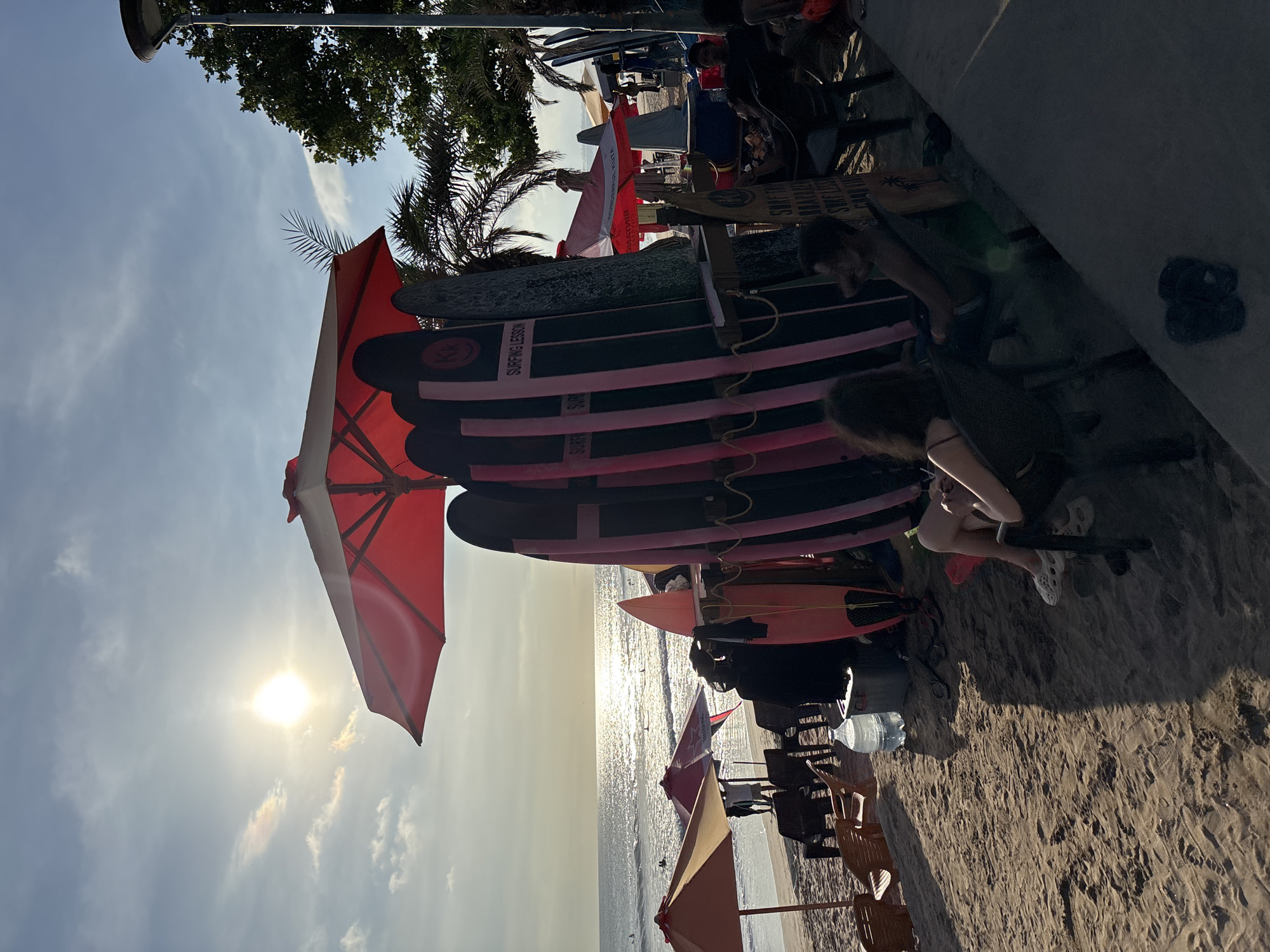 This is an outdoor shot taken in Kuta, Indonesia, likely at a beach. The sun glares off the water, indicating either sunrise or sunset. There are surfboards, some labeled "Surfing Lesson," stacked under a red beach umbrella. People are sitting nearby on chairs. Beach umbrellas and other seating arrangements are visible along the shoreline. The environment includes sand, palm trees, and a glimpse of buildings or structures along the upper edge of the scene.