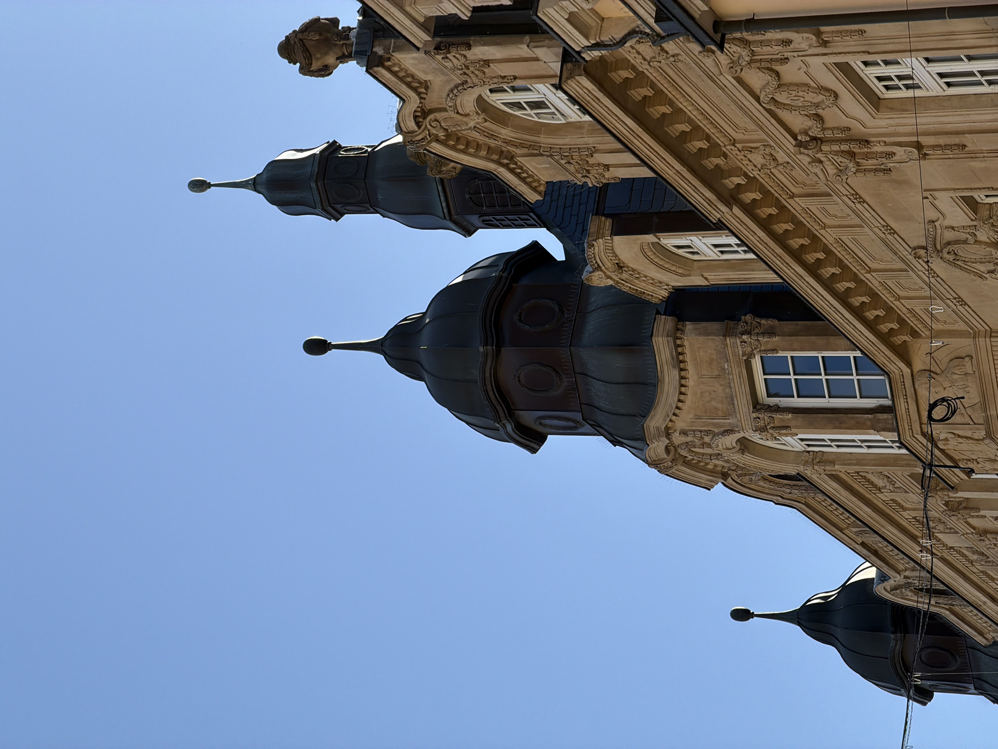The image presents a low-angle view of an ornate, historic building set against a clear, bright blue sky, suggesting a sunny day. The primary subjects are the architectural details of the building, characterized by intricate design and craftsmanship.
The building facade is a light, warm stone or stucco color, richly decorated with classical elements such as sculpted friezes, cornices, and decorative window surrounds. Several windows are visible, some rectangular with multiple panes, others featuring arched or circular designs.
Prominently featured are three dark, dome-shaped spires or turrets, two in the midground and one partially visible on the far right. These domes are segmented, tapering to a point, and topped with spherical finials. Their dark, possibly oxidized metal, surface contrasts sharply with the lighter building material and the vibrant sky.
A small, carved stone bust or sculptural element is visible near the top right edge of the building, adding to its grand aesthetic. A few dark cables or wires are also present, running vertically along the building's side, hinting at modern utilities integrated with the historic structure.
The scene captures a static architectural beauty, devoid of people or discernible activity. The cloudless blue sky dominates the background, reinforcing the bright daylight conditions. No visible text is present in the image. The building is located in Esch-sur-Alzette, Luxembourg.