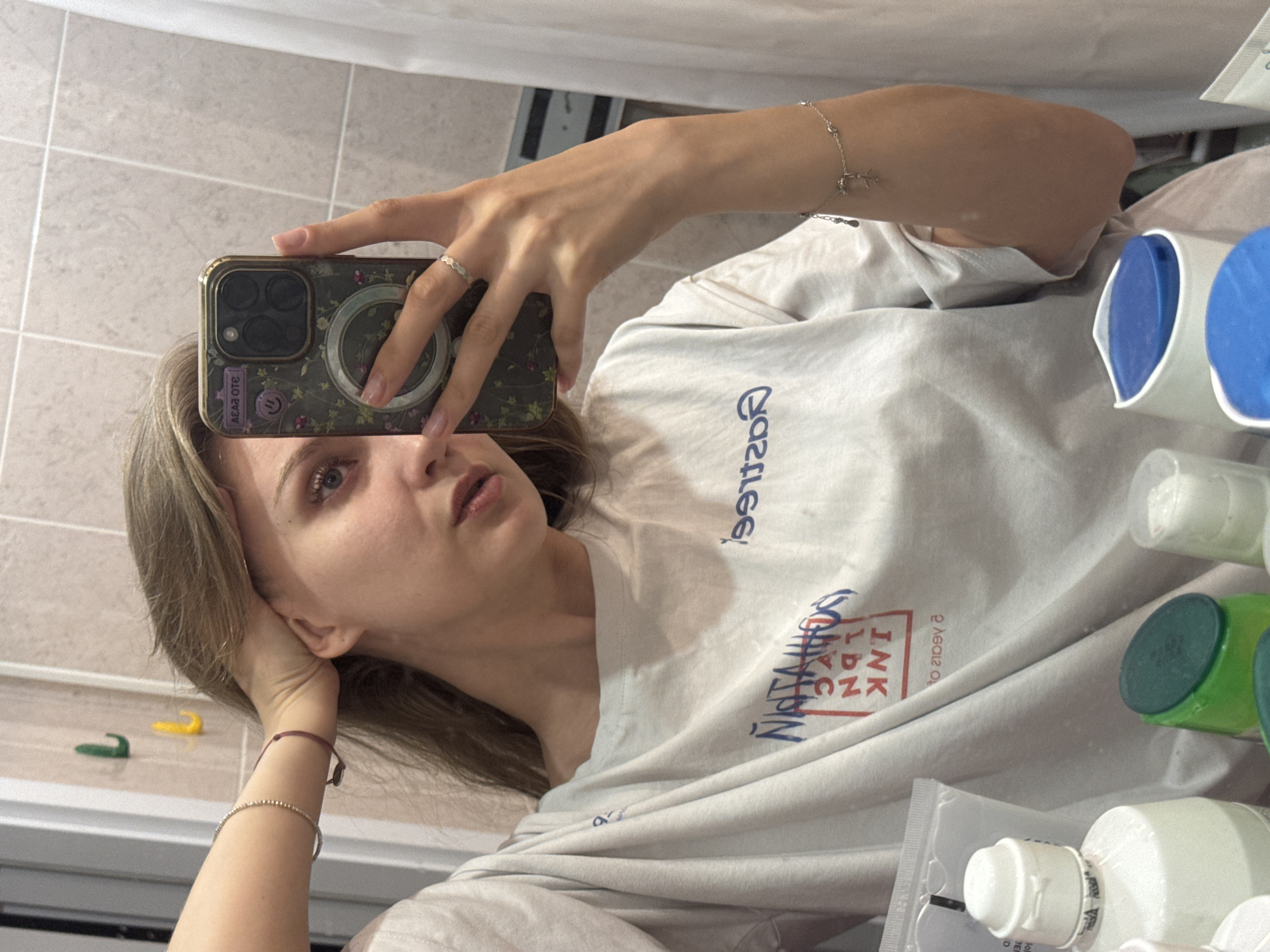 A young woman is taking a selfie in what appears to be a bathroom. She is holding her phone, which has a floral phone case, with her right hand. She is wearing a silver ring and a bracelet on her right wrist. She is wearing a white T-shirt with blue and red lettering, that says "Nastasia INK LDN NYC". Her left hand is resting on her head. There are bathroom supplies like tubes and bottles partially visible on the right side of the frame. In the background, there are tiled walls. The location is possibly a private residence. The image does not provide enough information to pinpoint a location within Izhevsk.