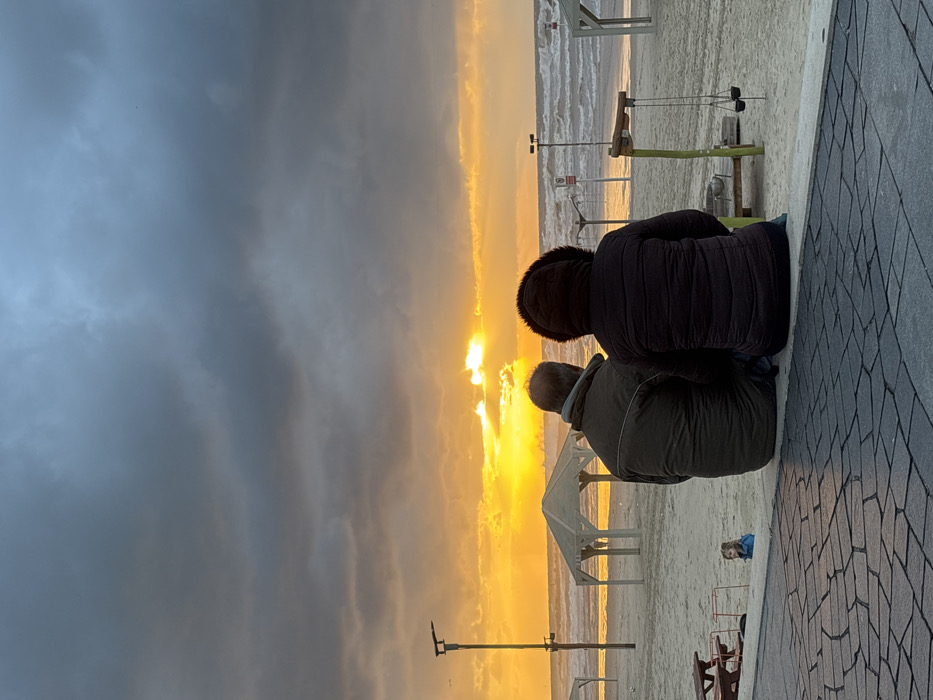 Two people sit together on a paved walkway, gazing at a vibrant sunset over the sea. Dark clouds loom on the left, contrasting with the bright orange and yellow hues reflecting off the water. Beach infrastructure, including lifeguard towers and benches, are visible in the distance. The figures are silhouetted against the bright sky, wearing warm jackets.