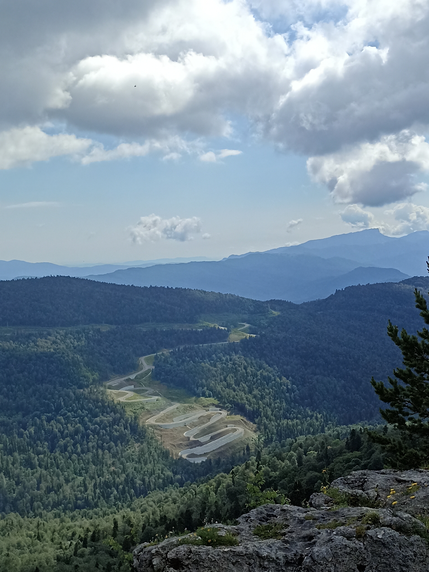 The image depicts a panoramic view of a mountainous landscape near Podgorny, Russia. In the foreground, a rocky outcrop with sparse vegetation and small yellow flowers is visible. A winding paved road, appearing as a light-colored ribbon, snakes its way down a steep, densely forested mountainside. The road exhibits numerous sharp turns and switchbacks, suggesting a significant elevation change. The surrounding slopes are covered with lush green trees, transitioning to darker hues as they recede into the distance. In the background, several layers of hazy blue mountains are discernible, fading into the horizon under a sky filled with a mix of white and gray clouds. A small, dark object, possibly a bird or an insect, is visible in the sky. The overall scene conveys a sense of vastness and natural beauty.