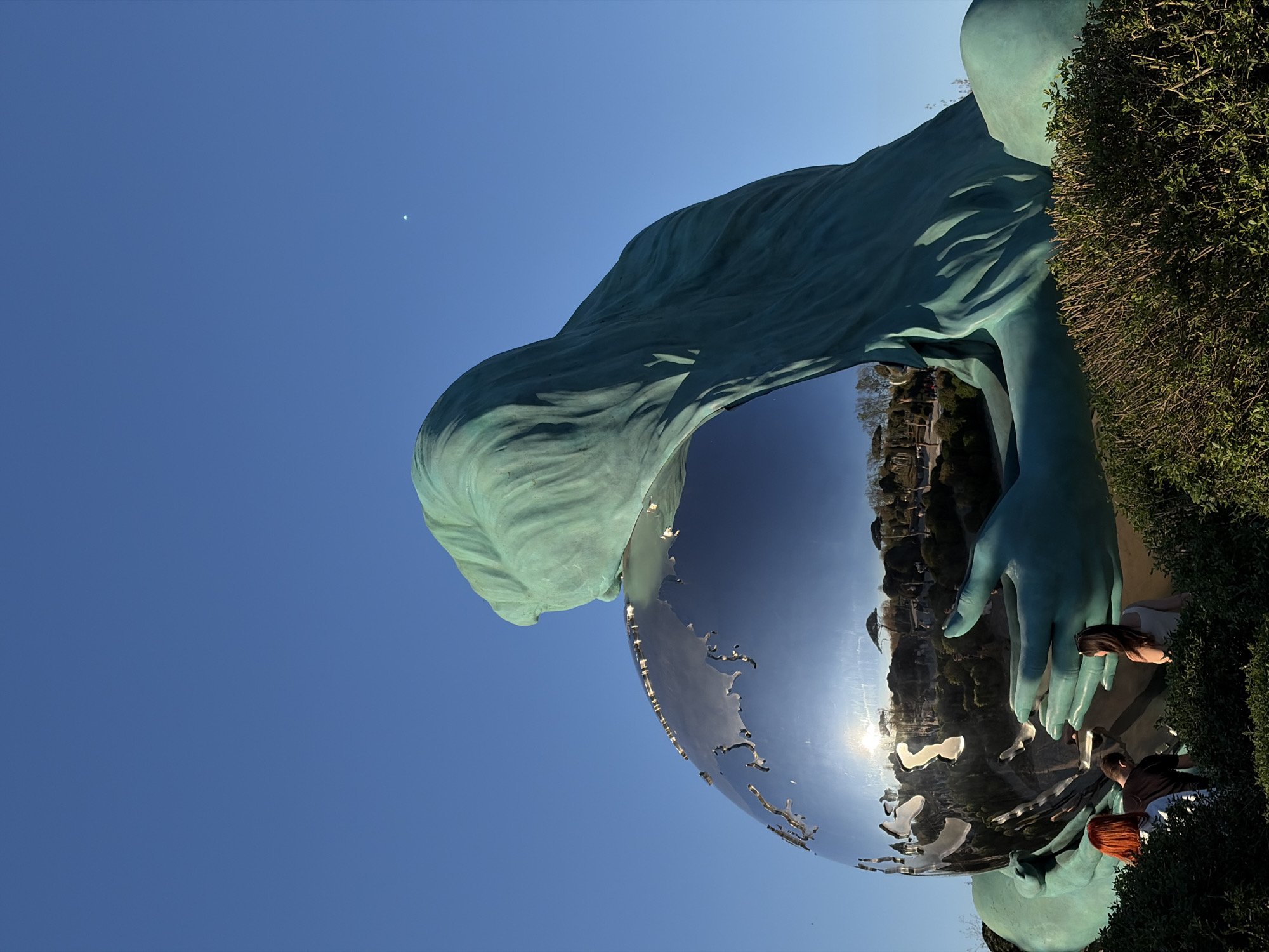 The image captures the "Reaching for the Sky" monument, a notable sculpture in the city of Krasnodar, Russia. The sculpture depicts a woman in teal holding a reflective chrome sphere resembling the Earth. Her hair flows down to touch the sphere. The sculpture is positioned beside a dense green hedge. Reflected in the sphere are buildings, vegetation, and three people. A woman with long brown hair is looking at the sphere. A boy in a dark shirt and a girl with red hair are crouched near the sphere, interacting with it. The background is a clear, light blue sky with a visible crescent moon in the upper left corner.