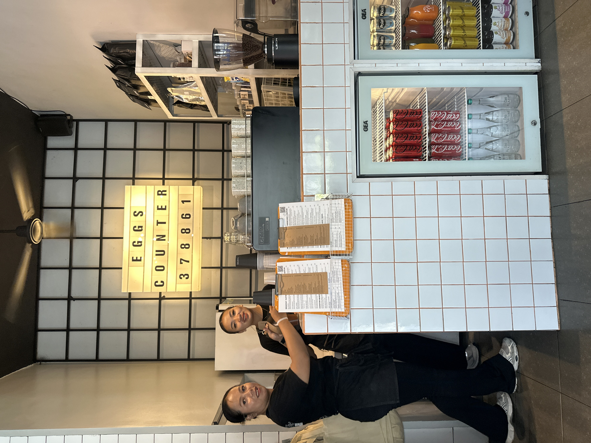 Two women, appearing to be staff members, are standing behind a counter in what seems to be a cafe or restaurant. The woman on the left is wearing a black t-shirt and black apron, while the woman on the right is wearing a black t-shirt. They are both looking towards the camera. In front of them are menus displayed on clipboards. Behind them, a lit sign reads "EGGS COUNTER 378861". The counter is made of white tiled material. To the right of the counter are two refrigerators displaying beverages, including cans of Coca-Cola and other drinks. The environment appears to be an indoor commercial establishment.