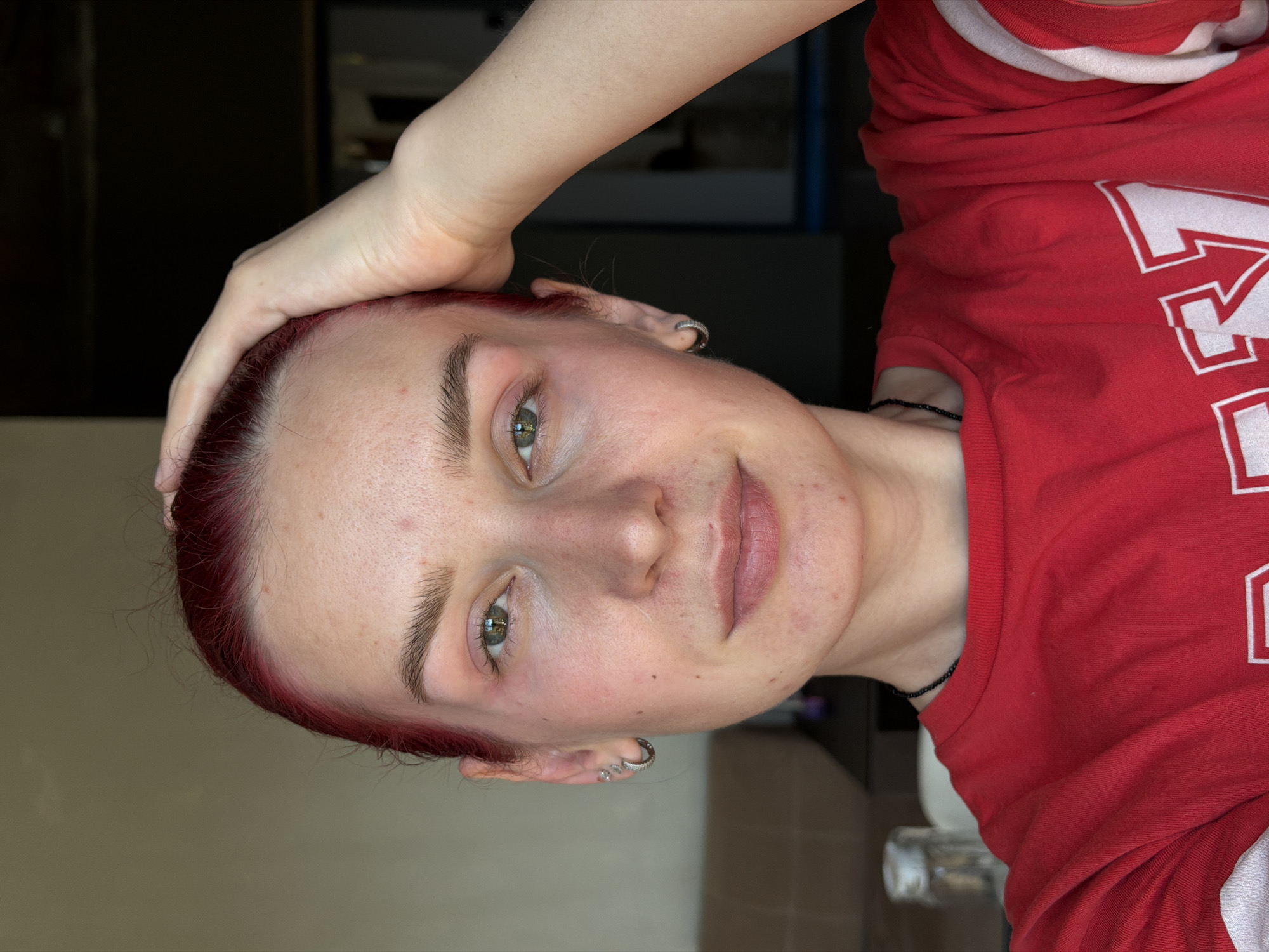A medium close-up shot captures a young woman with vibrant red hair, her right hand resting on the back of her head. She wears a red t-shirt with white accents. Her gaze is directed towards the camera, with one eye slightly more visible than the other. The background appears to be an indoor setting with soft lighting and a muted wall, suggesting a domestic environment. The image lacks any visual elements that would indicate the city of Chaikovsky, Russia, or any specific urban context.