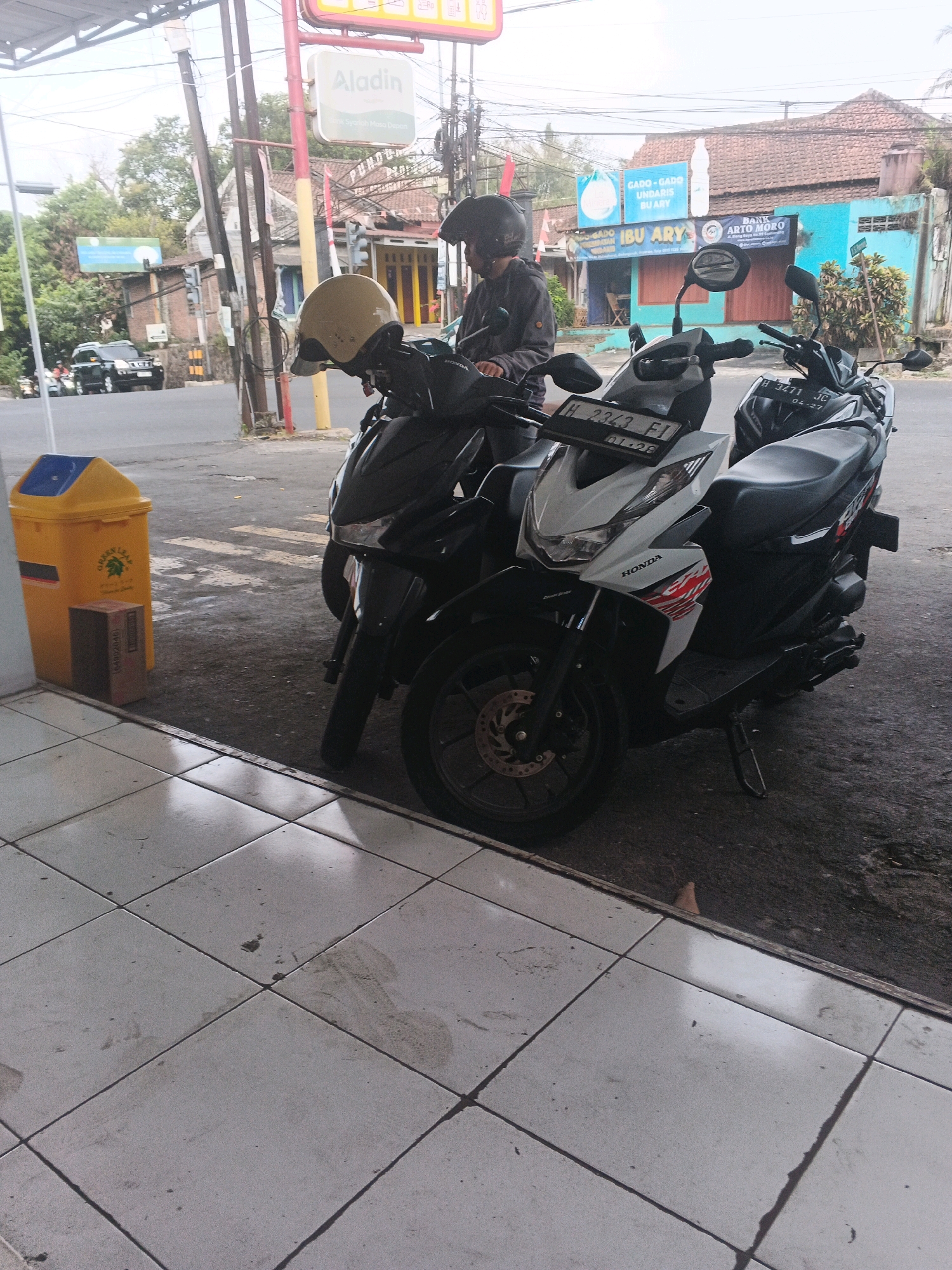 In the city of Sidomulyo, Indonesia, two motorcycles are parked side-by-side on a paved surface in front of a building. The motorcycle on the left is black, and the one on the right is white with red and black accents. A person wearing a black helmet and dark clothing is seated on the black motorcycle, facing away from the camera. The background shows a street with vehicles, buildings, and trees. A yellow trash can and a cardboard box are visible to the left of the motorcycles. The ground in the foreground is tiled.