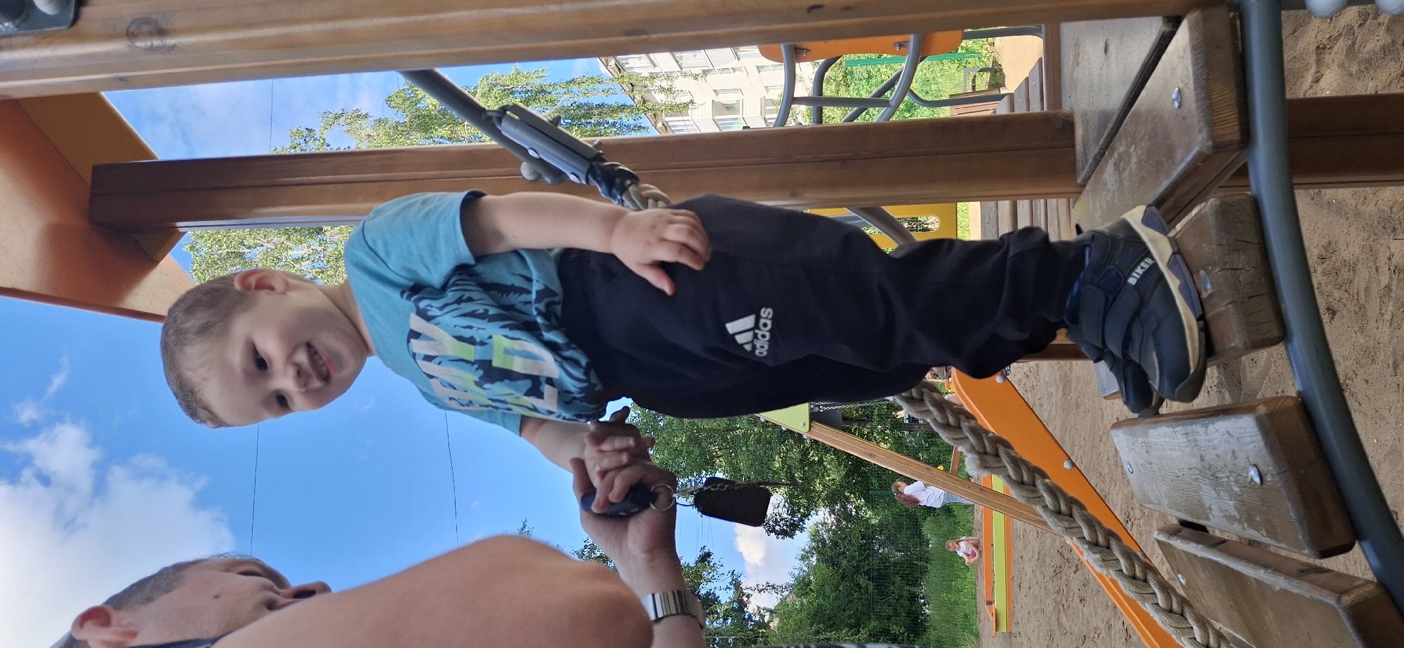 The image shows a young child, likely a toddler, suspended from a playground climbing structure. The child is wearing a blue t-shirt with a light blue, green, and white graphic pattern and black Adidas pants. The child's left hand is visible, gripping a chain or rope. The child's face is turned towards the camera, and they appear to be smiling.
Below the child, another person's arm and hand are visible, wearing a watch. This person seems to be holding onto the child or the climbing structure. The background reveals a sandy playground area with colorful climbing equipment made of wood and orange plastic. Green trees and a bright blue sky with scattered clouds suggest it is daytime and the weather is fair. In the distance, a building with multiple windows can be seen, possibly an apartment building.
The image is oriented vertically, with the child appearing to be at the top. The overall impression is one of playfulness and interaction between the child and the adult. The text "adidas" is visible on the child's pants.