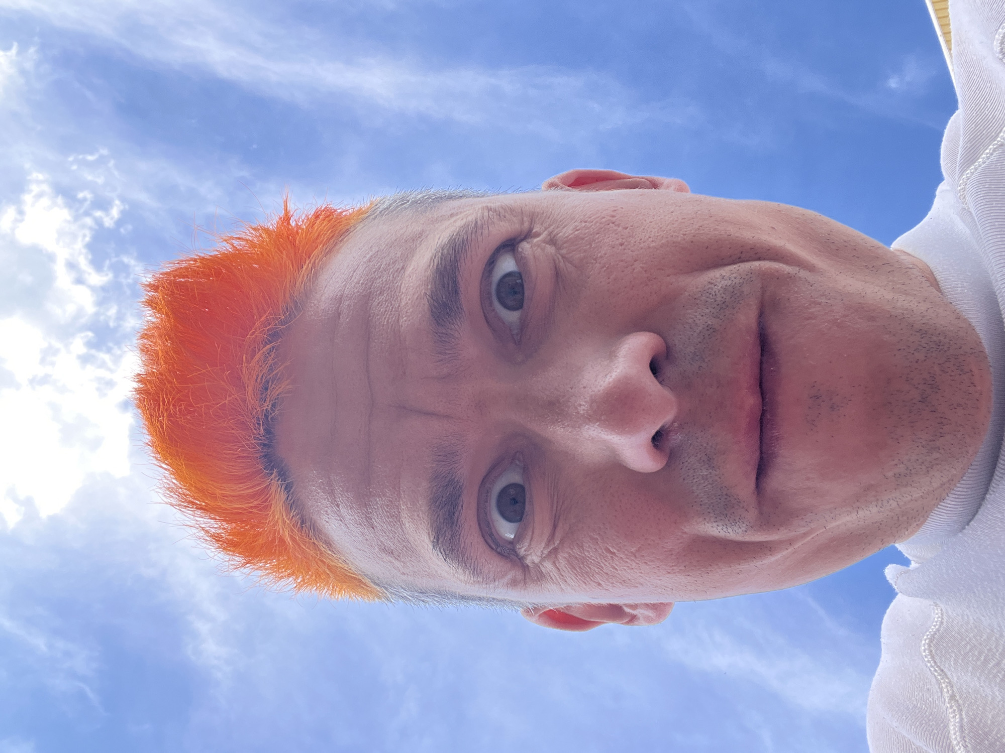 A selfie of a person with orange hair taken from a low angle against a sky with scattered clouds. The person has light skin, brown eyes, and some stubble. They are wearing a white shirt. The photograph was taken outdoors during daylight hours. There is no information available to determine the location as Chaikovsky, Russia.
