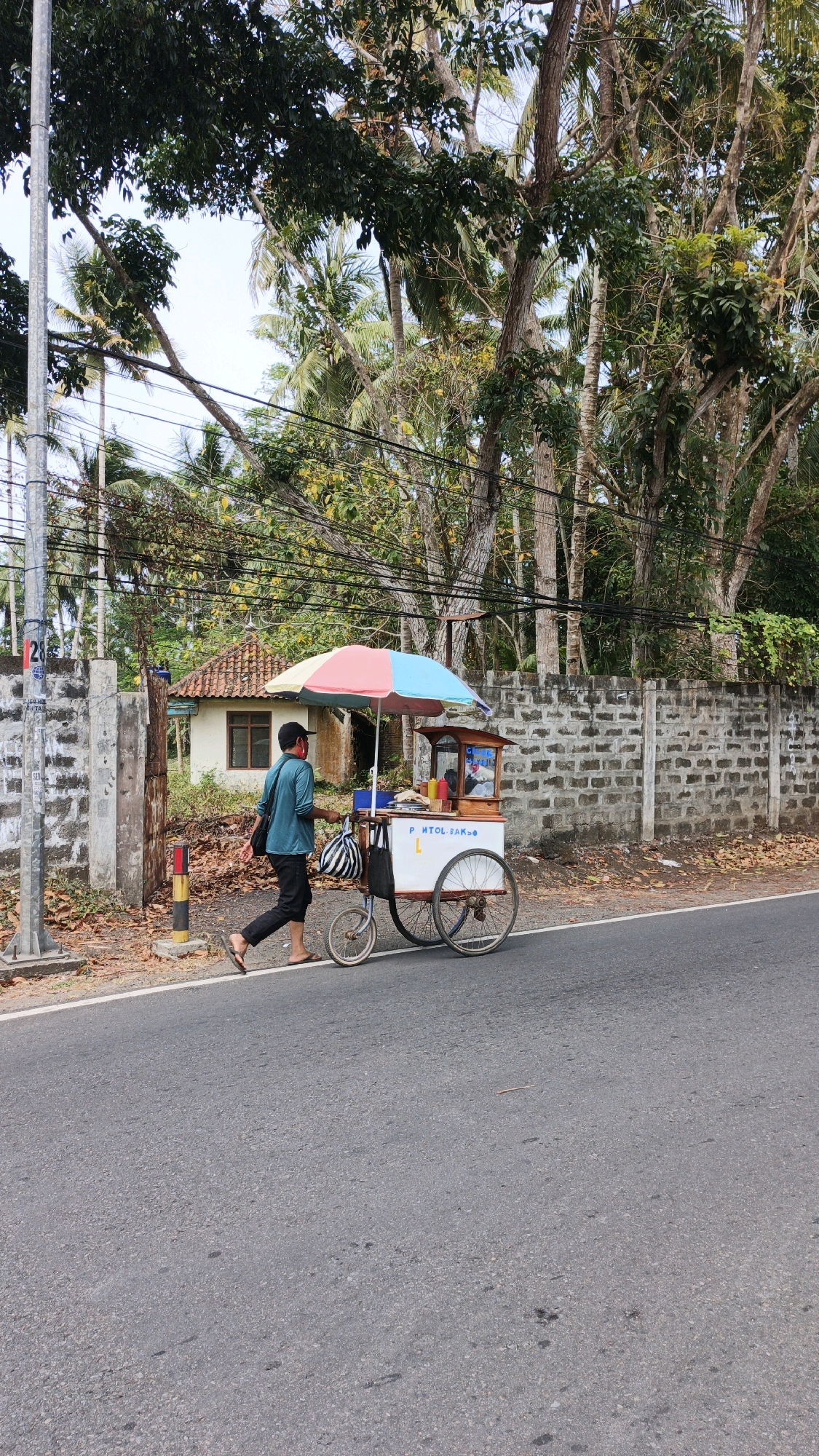 A man wearing a teal shirt, black pants, and flip-flops walks his food cart along the side of a paved road in Cibenda, Indonesia. He is wearing a red mask and carrying a black bag over his shoulder. The food cart has a colorful umbrella and displays bottles of condiments. Behind the man, a low brick wall borders a property with a small house and dense vegetation. Overhead, power lines are visible among the trees.