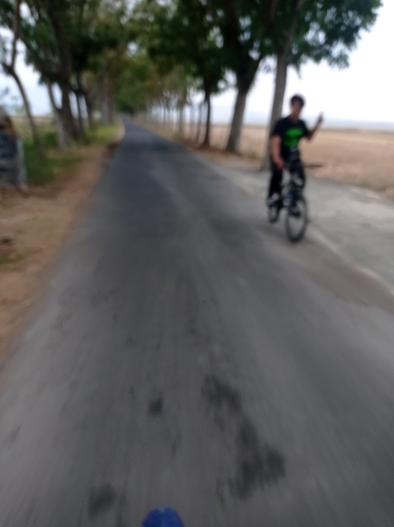 A person on a bicycle rides down a paved road lined with trees. The person is wearing a dark t-shirt with a green accent and dark pants. They are holding up their right hand, possibly waving. The road is gray and appears to be in a rural or suburban area, likely in Sumilir, Indonesia, given the foliage and landscape. The sky is overcast. The image is blurry and taken from a low angle, suggesting the photographer is also on a bicycle. There is a hint of dry grass or fields in the background.