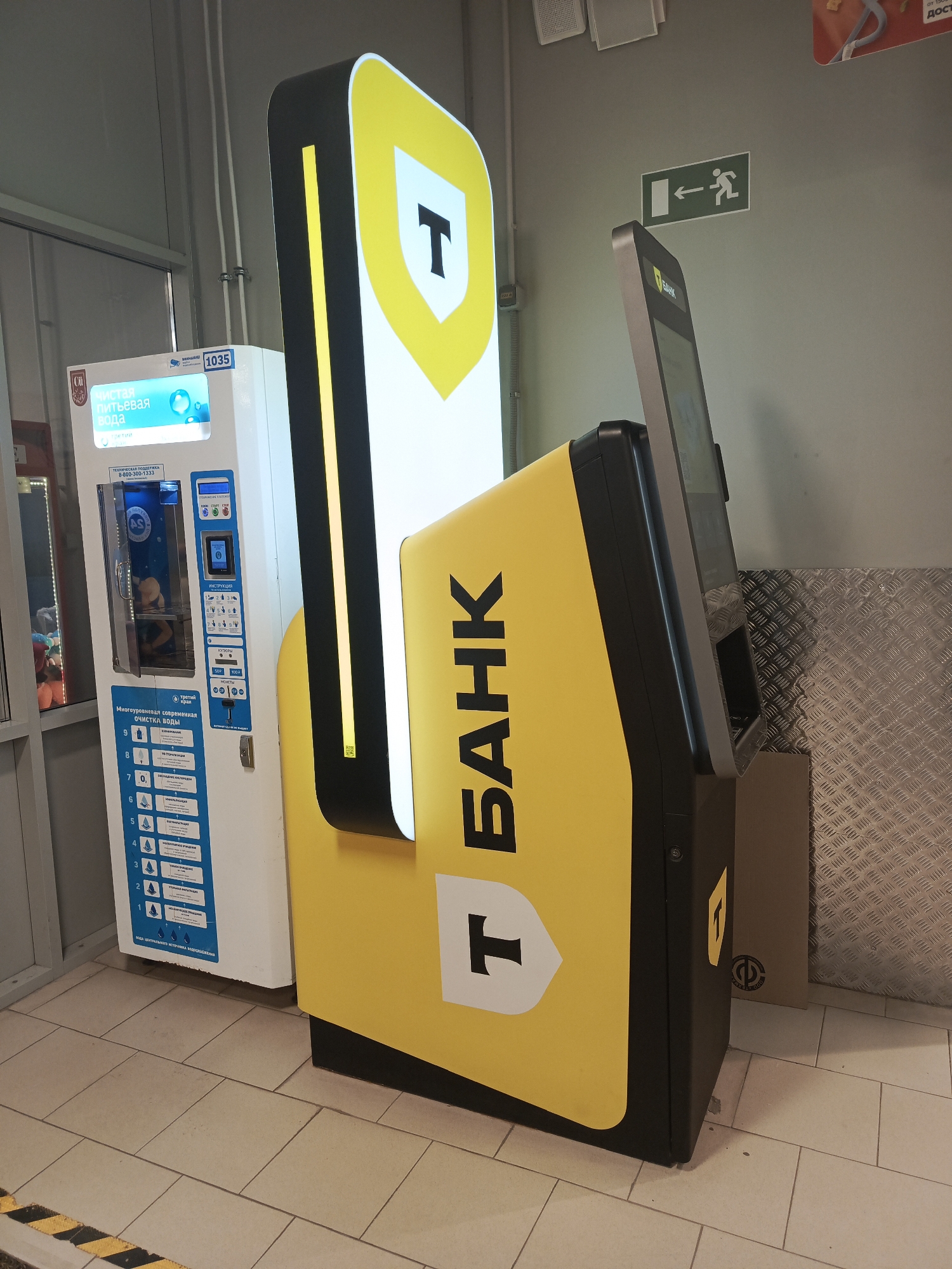 In Perm, Russia, a photograph captures a corner of a commercial or public space, likely an indoor facility. To the left of the frame, a white water vending machine is visible, displaying text in Russian, including "ЧИСТАЯ ПИТЬЕВАЯ ВОДА" (Clean drinking water) and instructions for use. The machine features a transparent door revealing internal components and a payment slot.
Dominating the center and right of the image is a large, black and yellow kiosk or ATM, branded with a prominent "T" logo within a shield, and the word "БАНК" (Bank) in large, bold letters. The kiosk has a touchscreen display. An illuminated yellow vertical accent runs along the left edge of the kiosk.
On the wall behind the kiosk, a green and white exit sign with an arrow indicating a rightward escape route is affixed. The environment features tiled flooring and a corrugated metal wall in the background on the right. The lighting appears to be artificial and diffused. No people are present in the immediate scene.