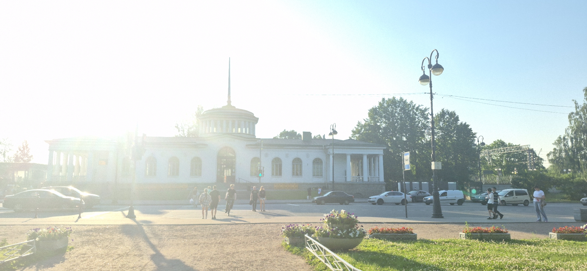 This image depicts a sun-drenched street scene in front of a white building with classical architectural elements, including columns and a domed roof structure. Several people are visible, some walking on the sidewalk and crossing a street. Cars and vans are also present, some parked and others driving. The scene is illuminated by bright sunlight, casting long shadows. In the foreground, there are planters with flowers and grassy areas. Trees and streetlights are also part of the environment. The overall impression is of a public space on a clear, sunny day.