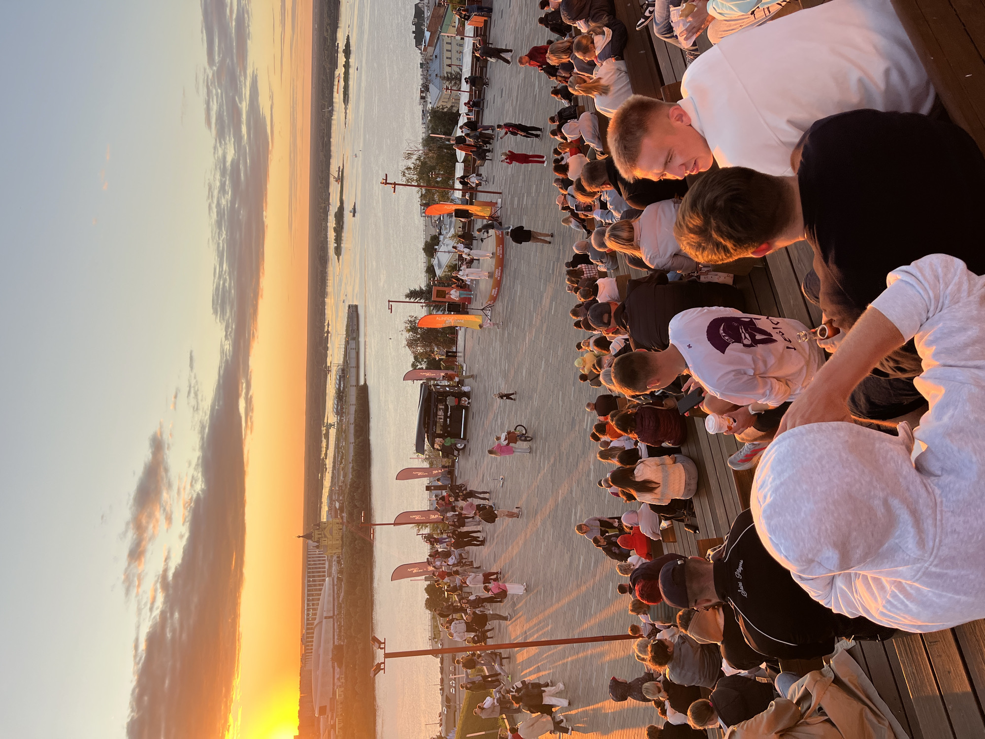 This is a vertical, high-angle, medium shot of a crowd of people gathered outdoors during what appears to be a sunset. The setting is a wooden promenade overlooking a wide body of water, likely a river or bay, with a city skyline visible in the distance on the far bank. The sky is awash with the warm oranges and yellows of sunset, with some wispy clouds.
The crowd is dense and appears to be seated on wooden benches or standing on a paved area. Many people are looking towards a small stage or performance area in the middle ground. On this stage, a small group of individuals dressed in white are performing, with several orange flags with text on them planted behind them. A black food truck or van is also present on the promenade. A few people are walking around, including a person on a bicycle. The overall mood seems relaxed and engaged, with people enjoying the outdoor event and the sunset.
Notable details include the warm, golden light of the sunset casting long shadows. The water is calm. The architecture of the distant city is visible, with some taller buildings and a prominent domed structure. The text on the orange flags appears to be in Cyrillic, consistent with a Russian location. One visible text fragment on the black jacket of a person in the foreground reads "Saint Players". The event could be a concert, festival, or public gathering, taking advantage of the pleasant evening weather.