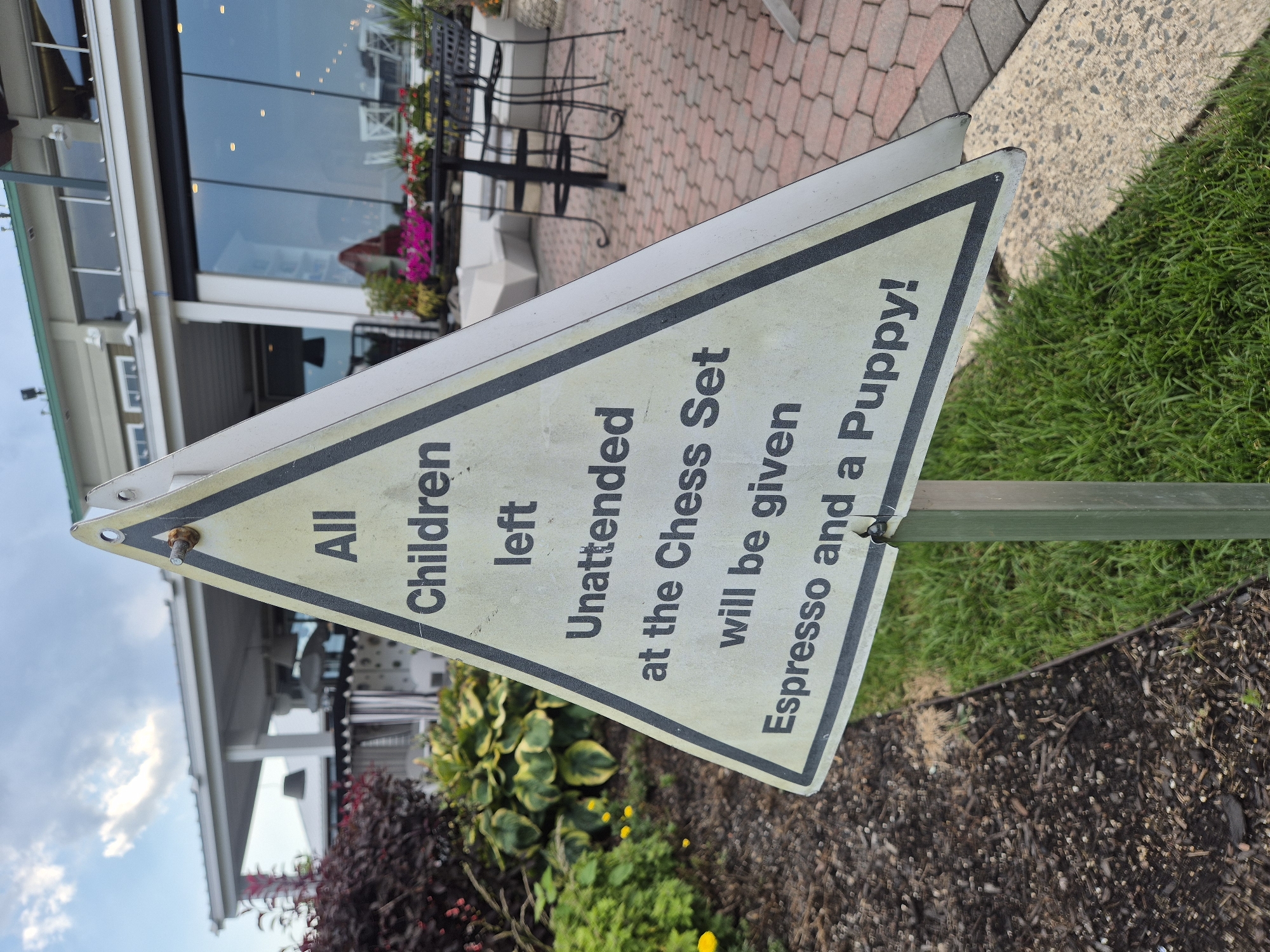 A triangular sign with black lettering on a cream-colored background is positioned in a garden area. The sign reads, "All Children left Unattended at the Chess Set will be given Espresso and a Puppy!". Behind the sign, a building with large windows and outdoor seating is visible. The building appears to be a restaurant or cafe. The foreground consists of mulch and grass. The scene suggests a playful warning at an outdoor recreational area.