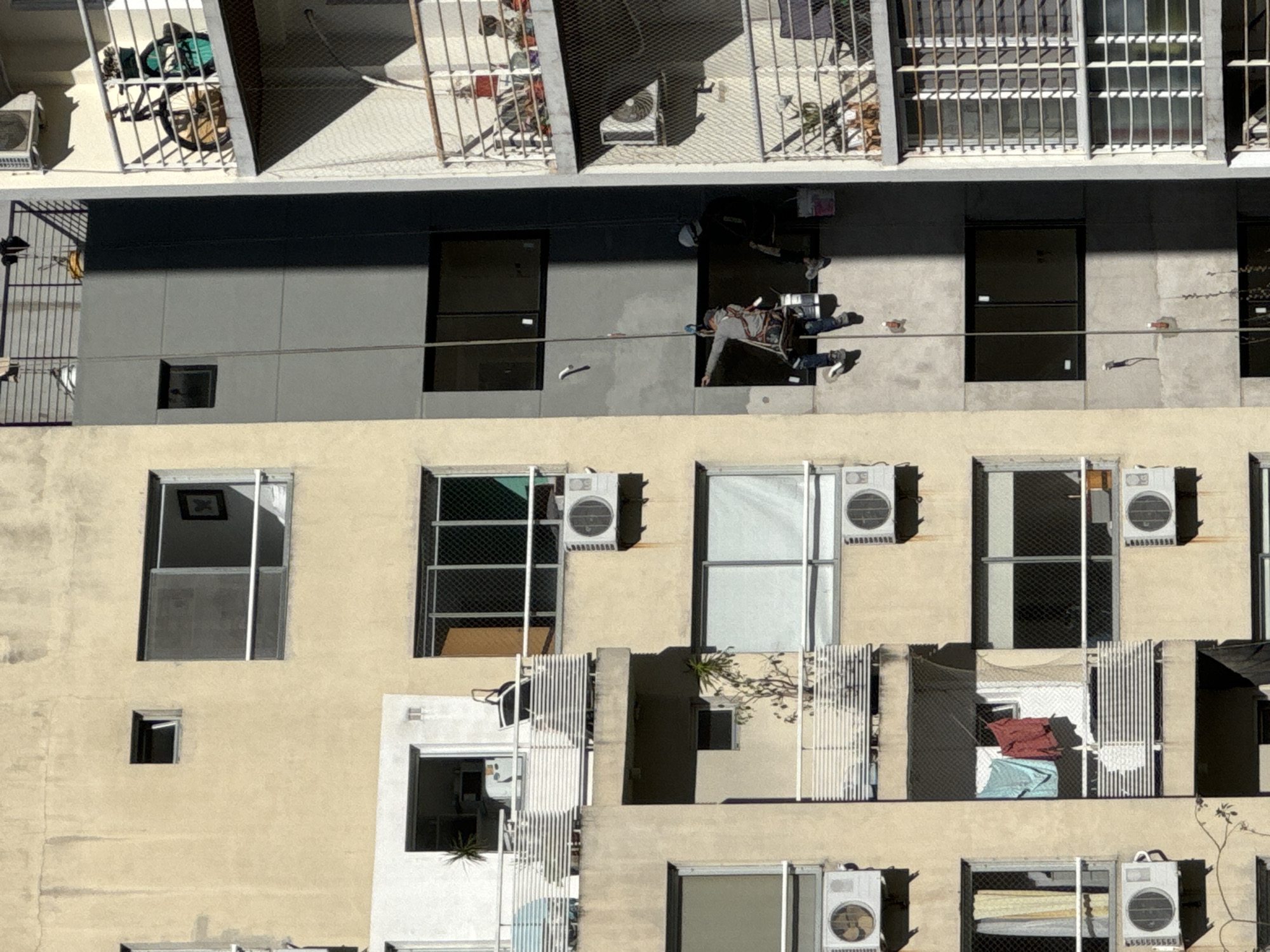 A high-angle, full shot shows the exterior of a building in Buenos Aires, Argentina. The building is a light beige color, with various windows and air conditioning units. A window washer is visible on the upper portion of the building. He is suspended by a rope and harness, working between two windows. The building's architectural design includes varying depths, creating light and shadow effects on the facade. Some windows have bars or safety grilles, and some are open, revealing interiors with clothes or other objects.